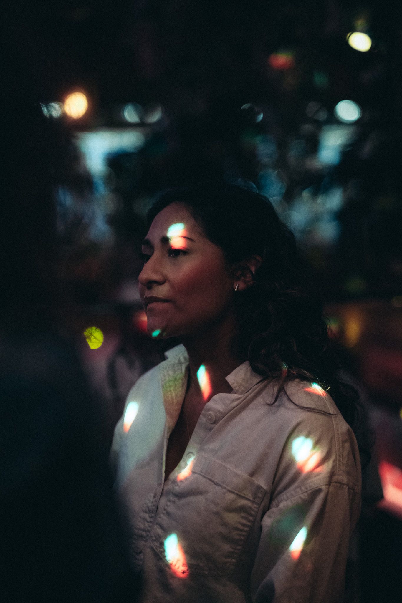 A woman in a light shirt stands in a dark outdoor setting illuminated by colorful bokeh lights.
