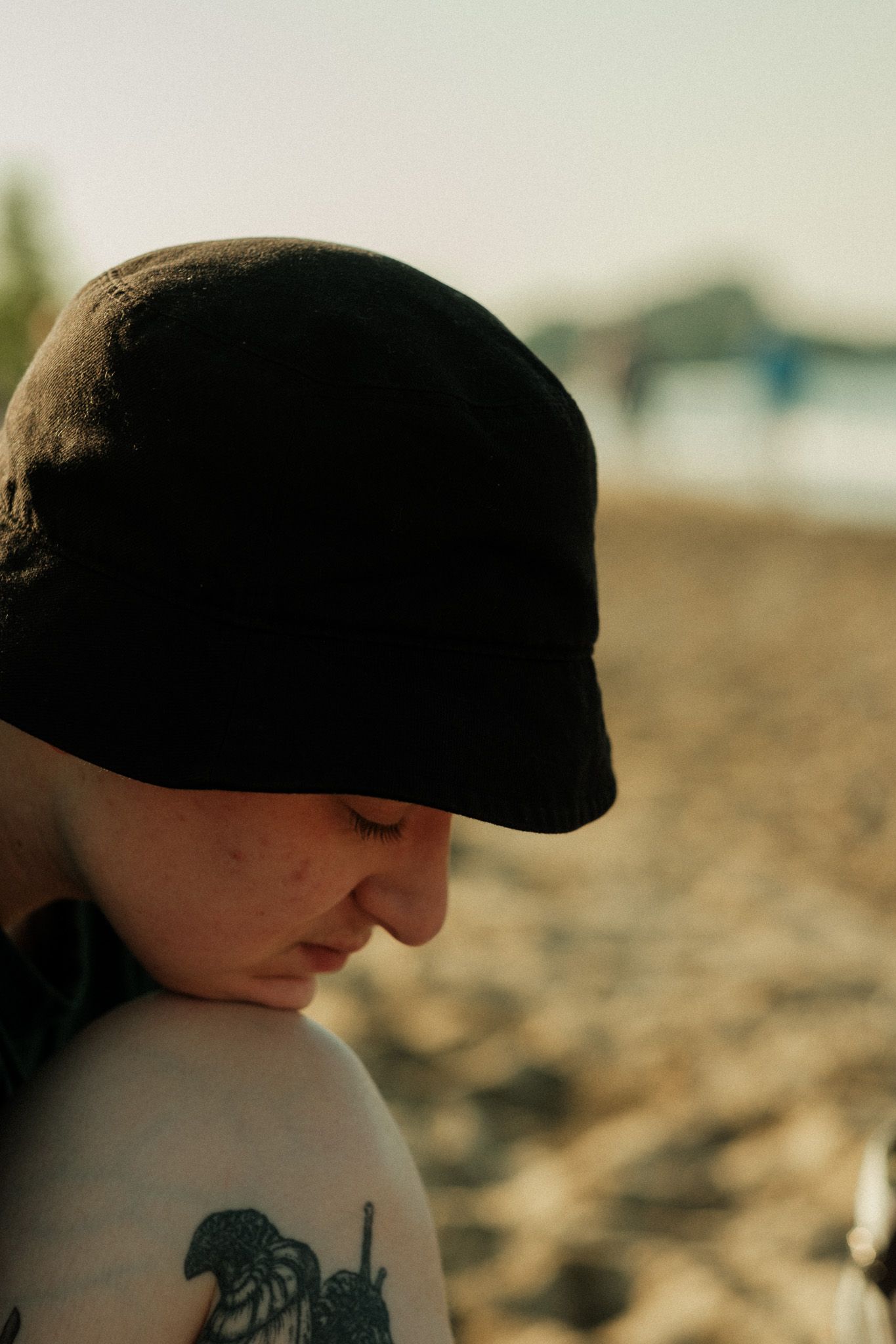 Close-up portrait of a person with a black bucket hat and floral tattoo on shoulder at beach.