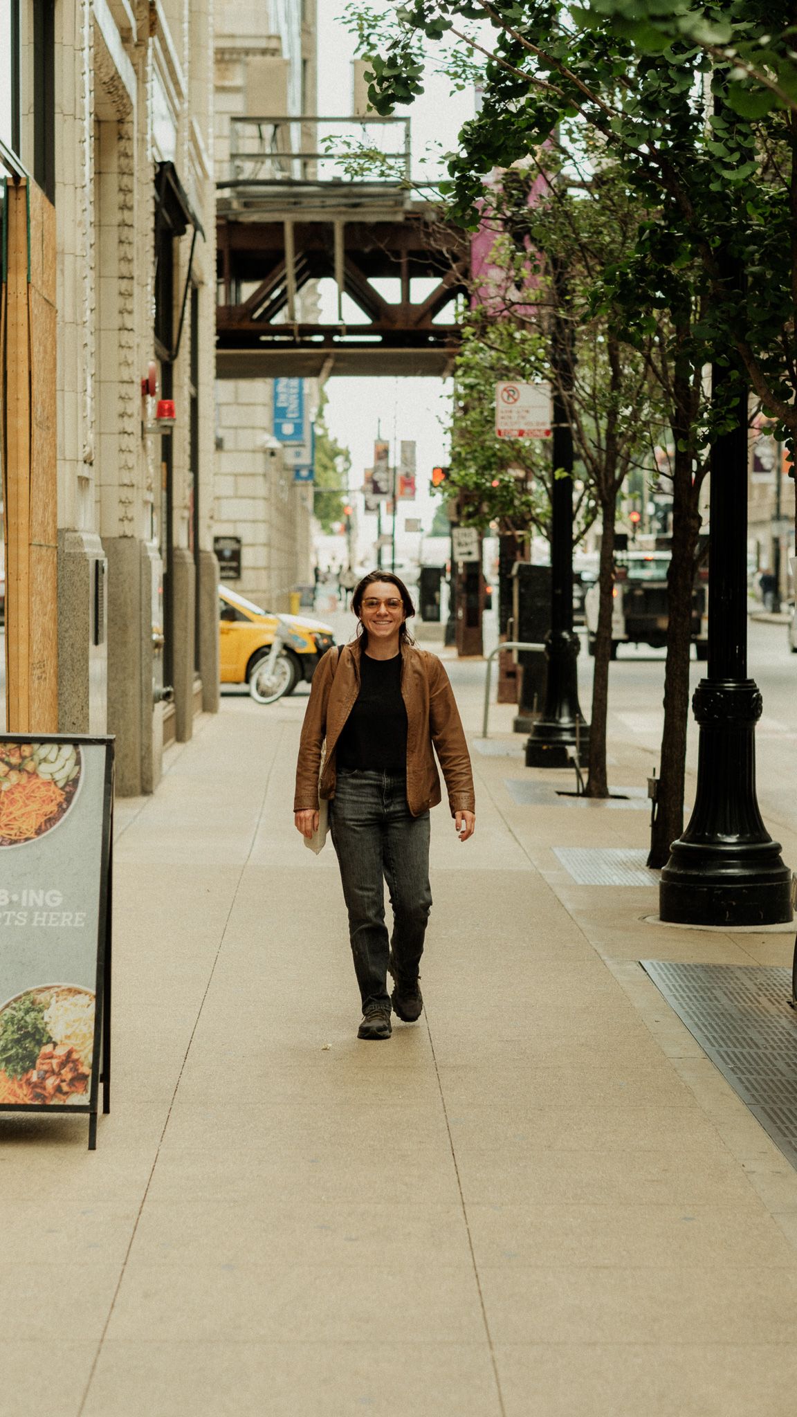 A person in a brown leather jacket walks confidently along a city sidewalk lined with trees.