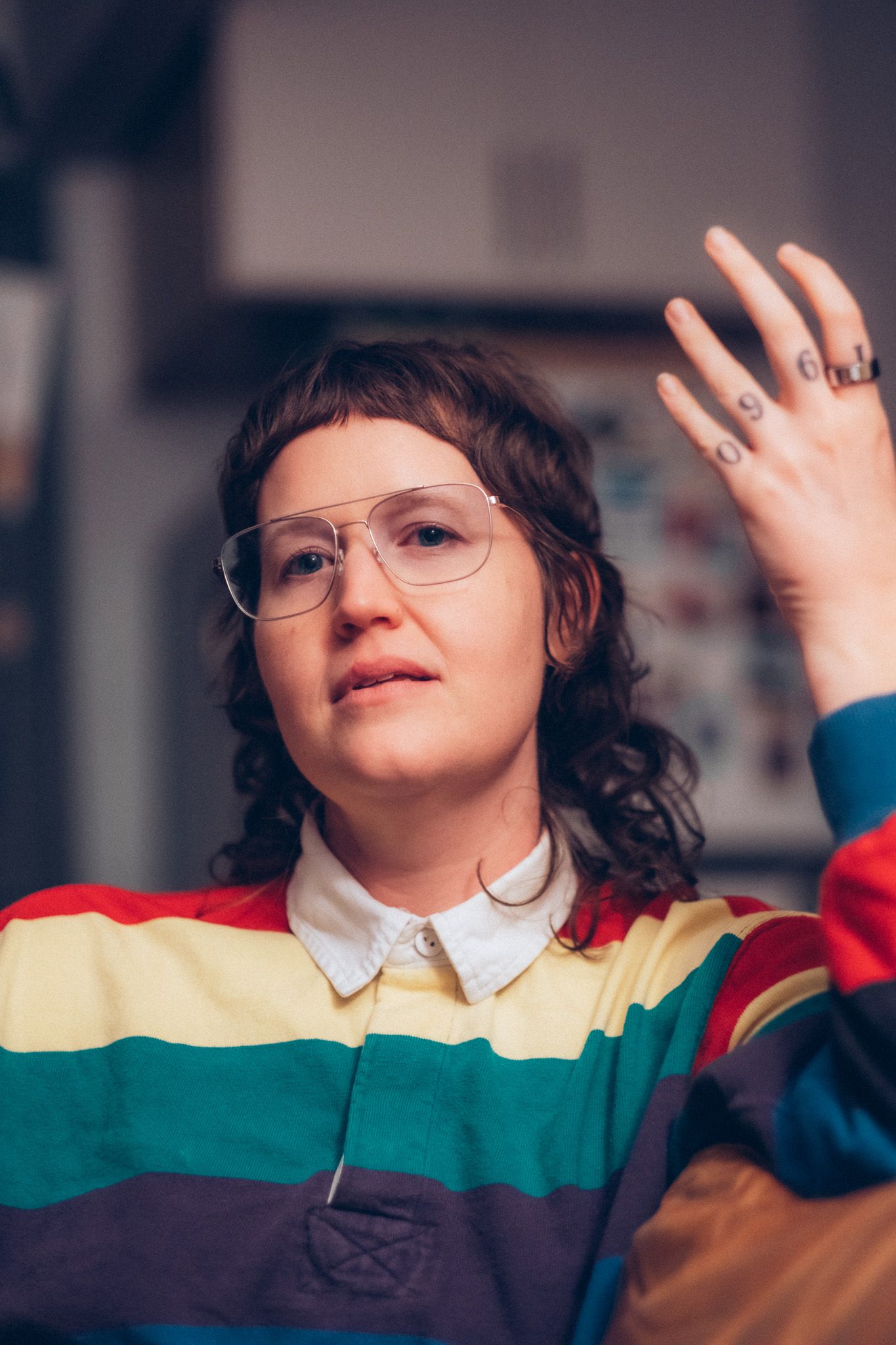 Young woman with curly hair wearing glasses and a colorful rainbow striped polo shirt, raising her tattooed hand.