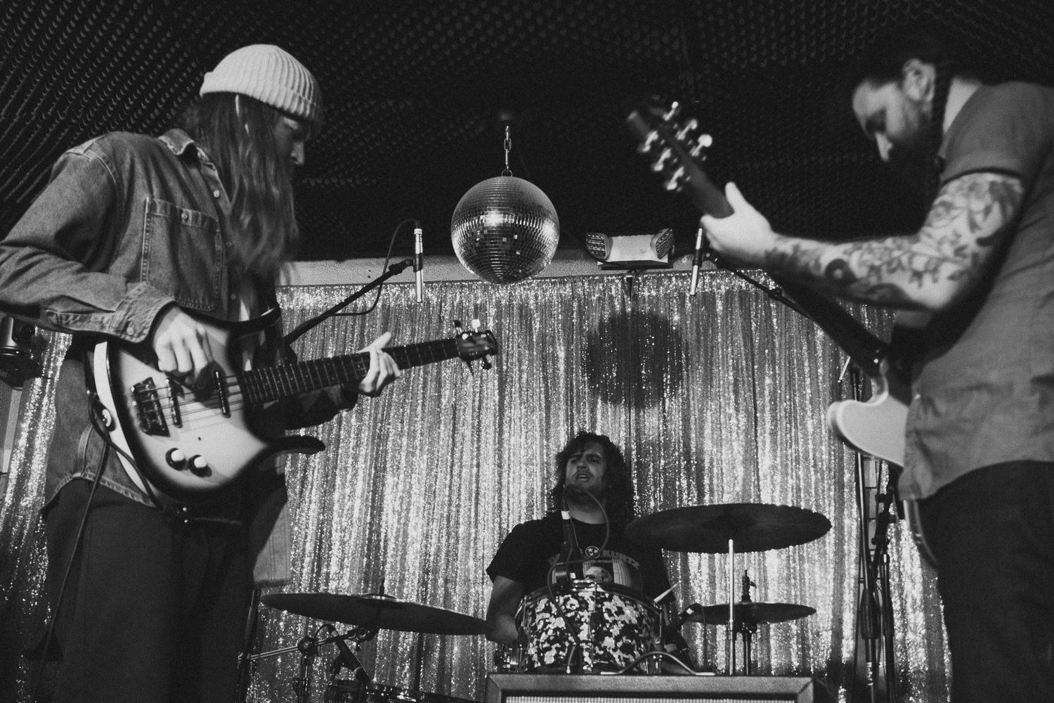 Black and white photo of a three-piece band performing on stage with a disco ball backdrop.