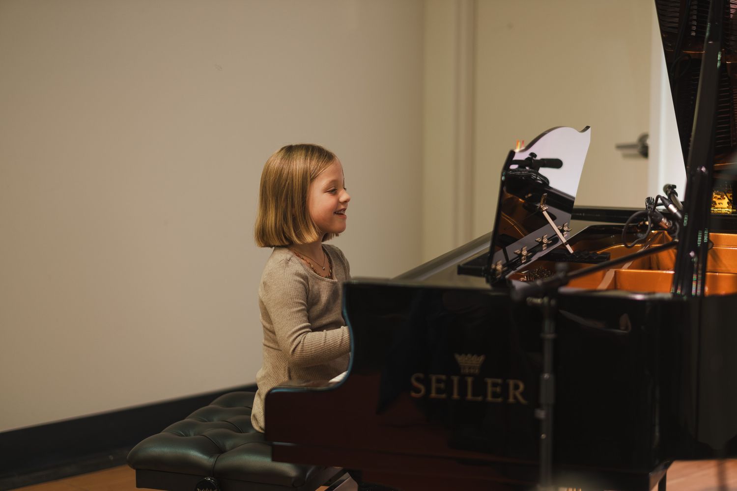 Young girl smiling while playing a Seiler grand piano during a music recital in a bright room.