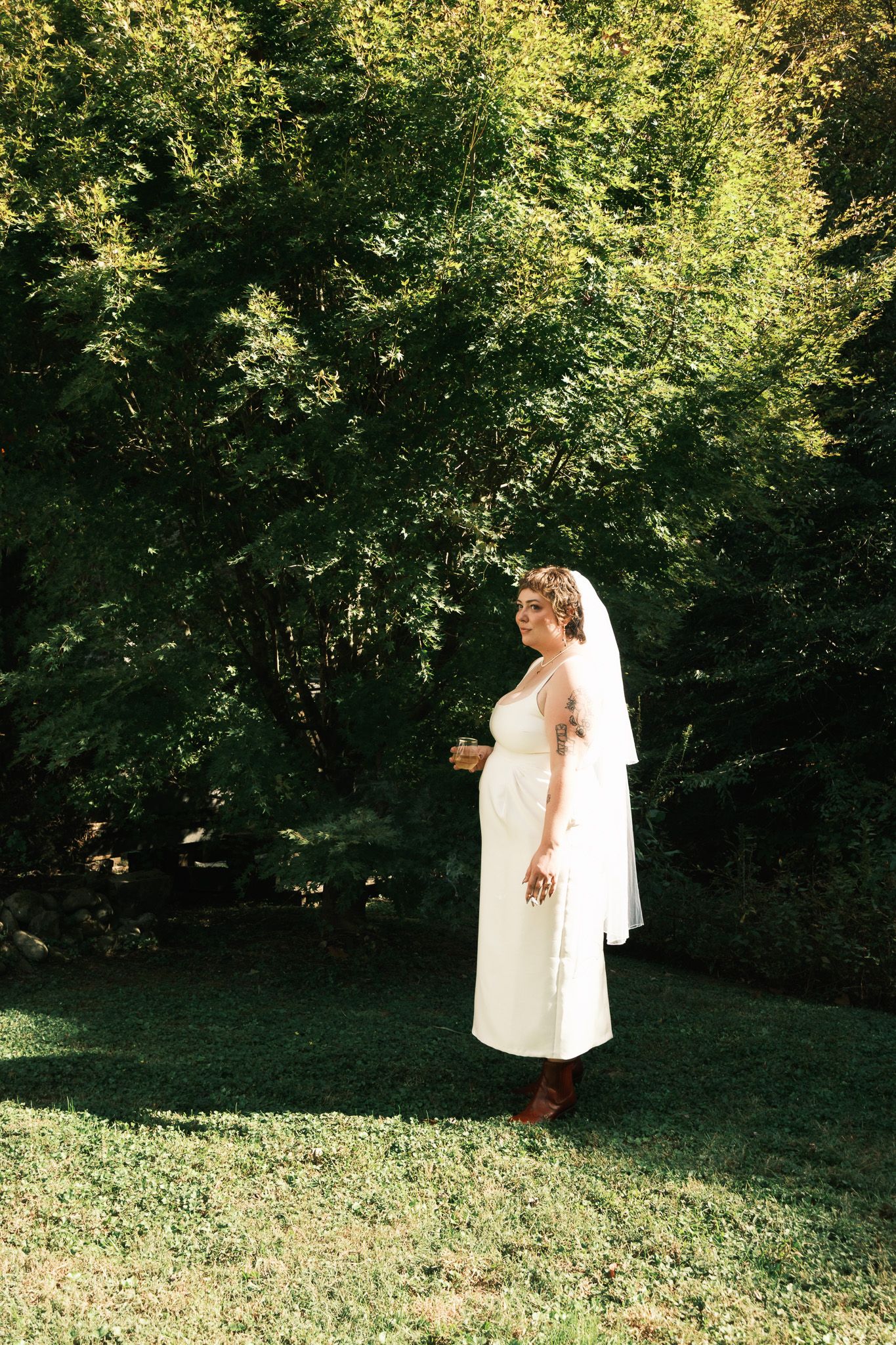 Bride in white midi dress and veil stands on green lawn surrounded by lush trees on her wedding day.