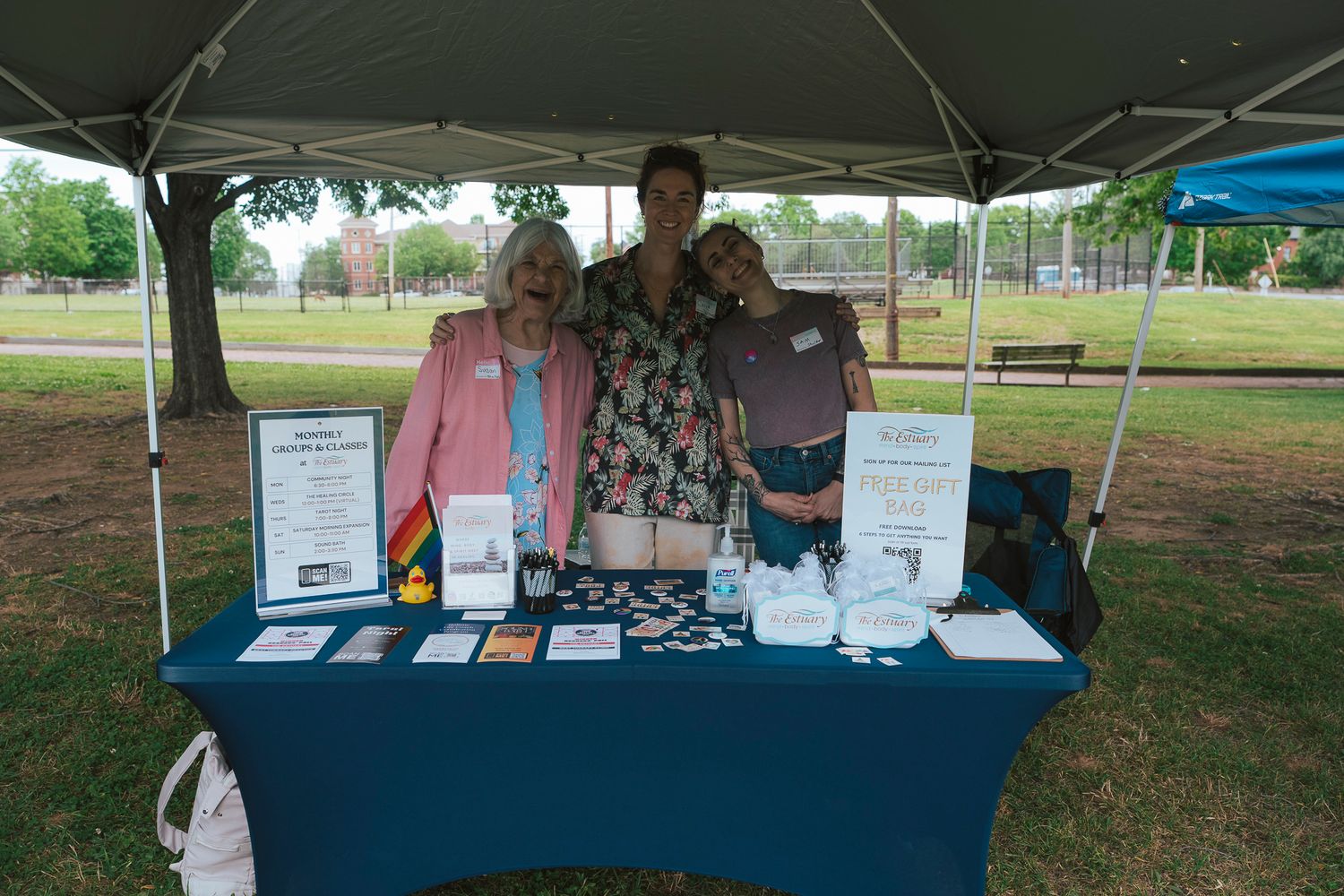 Three people posing together at an outdoor event booth with informational materials on a blue table.