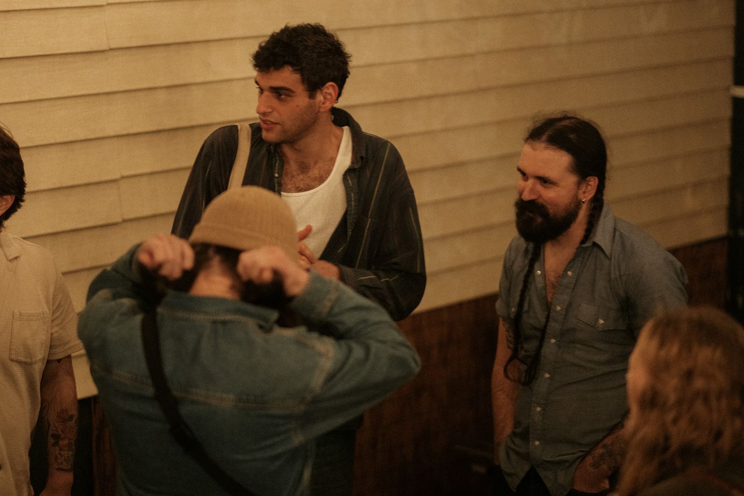 Three men stand in conversation near a wooden wall in a dimly lit indoor venue.
