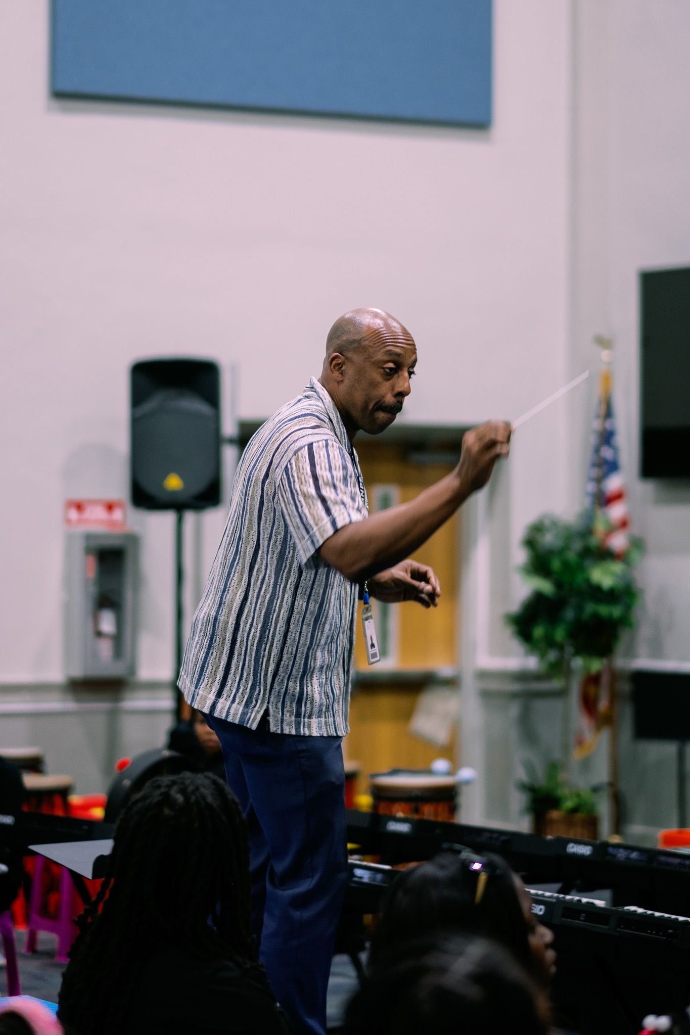 Male conductor holding a baton leads a student music ensemble in a school auditorium with an American flag.