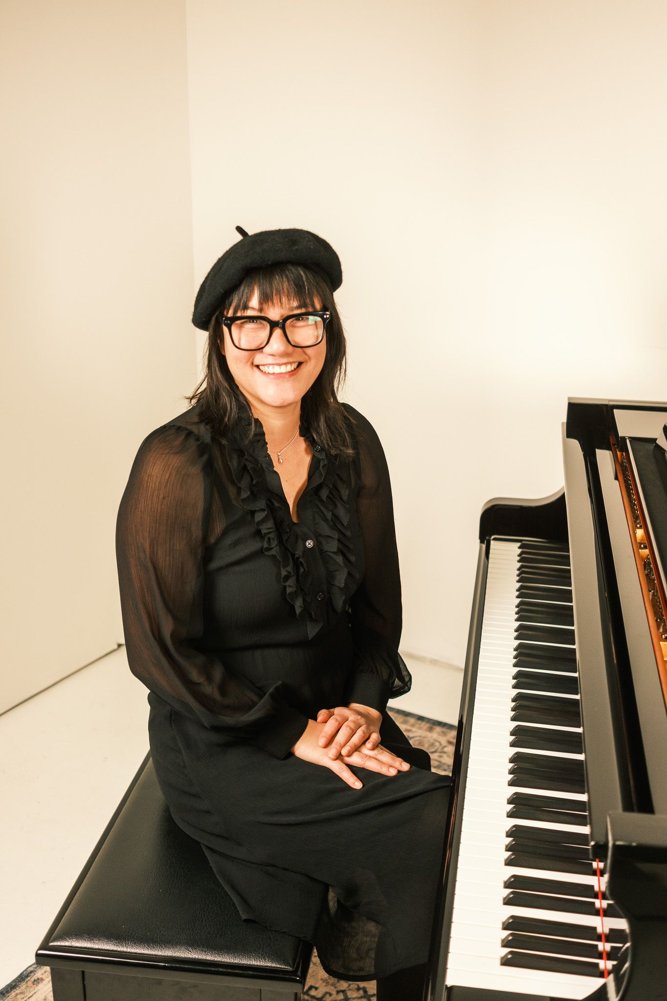 A smiling woman in a black beret and outfit sits beside a grand piano in a bright white studio.