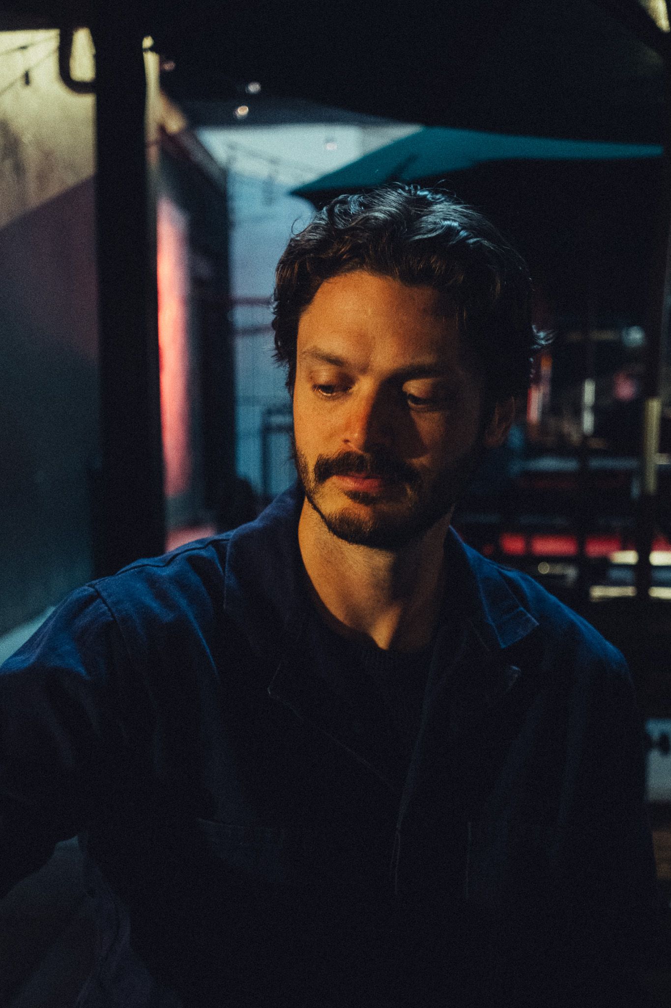 Bearded man with curly hair wearing a dark jacket, eyes closed, illuminated by warm moody lighting indoors.