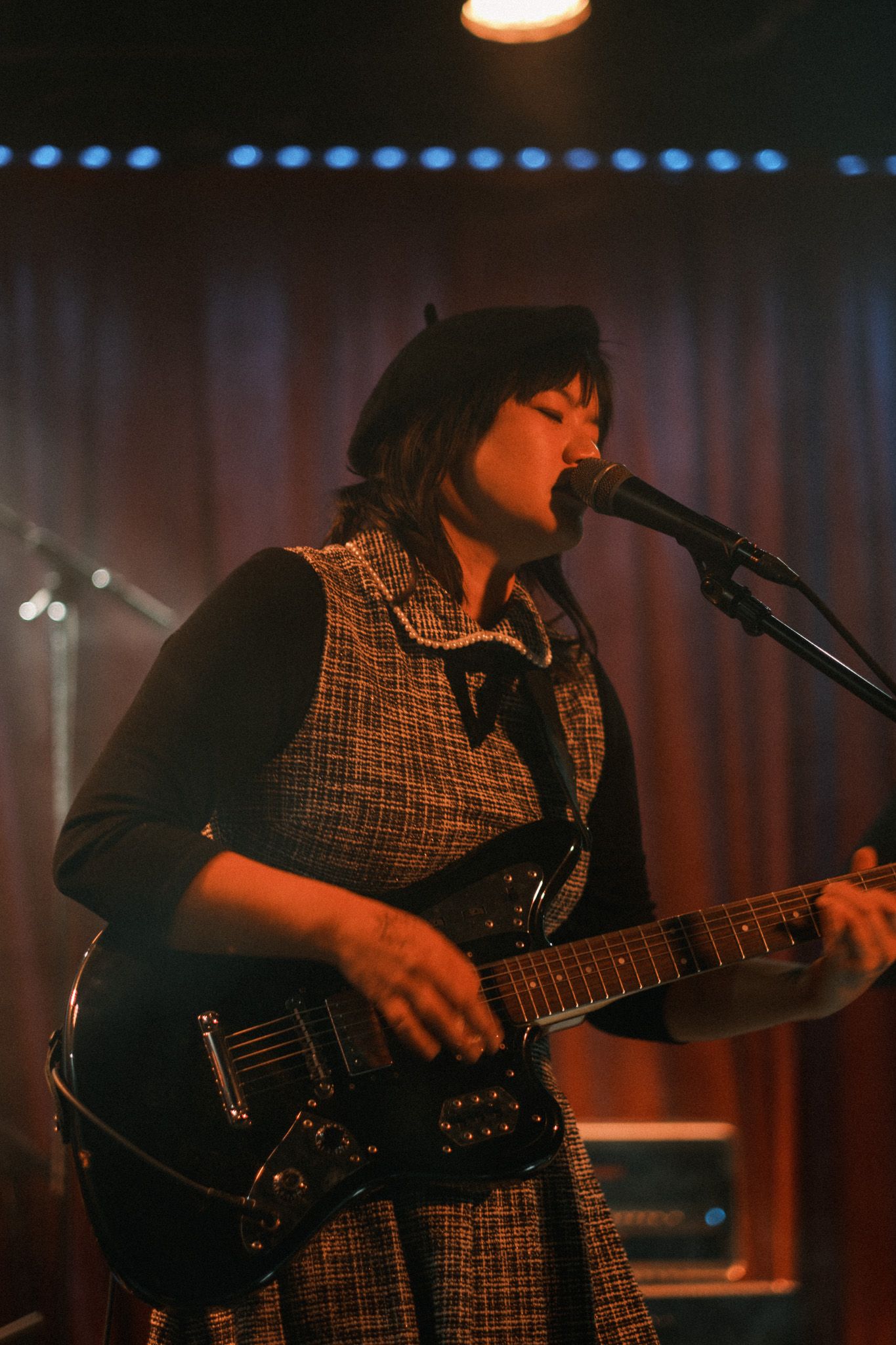 Female vocalist in beret and tweed vest playing electric guitar and singing into microphone on stage.