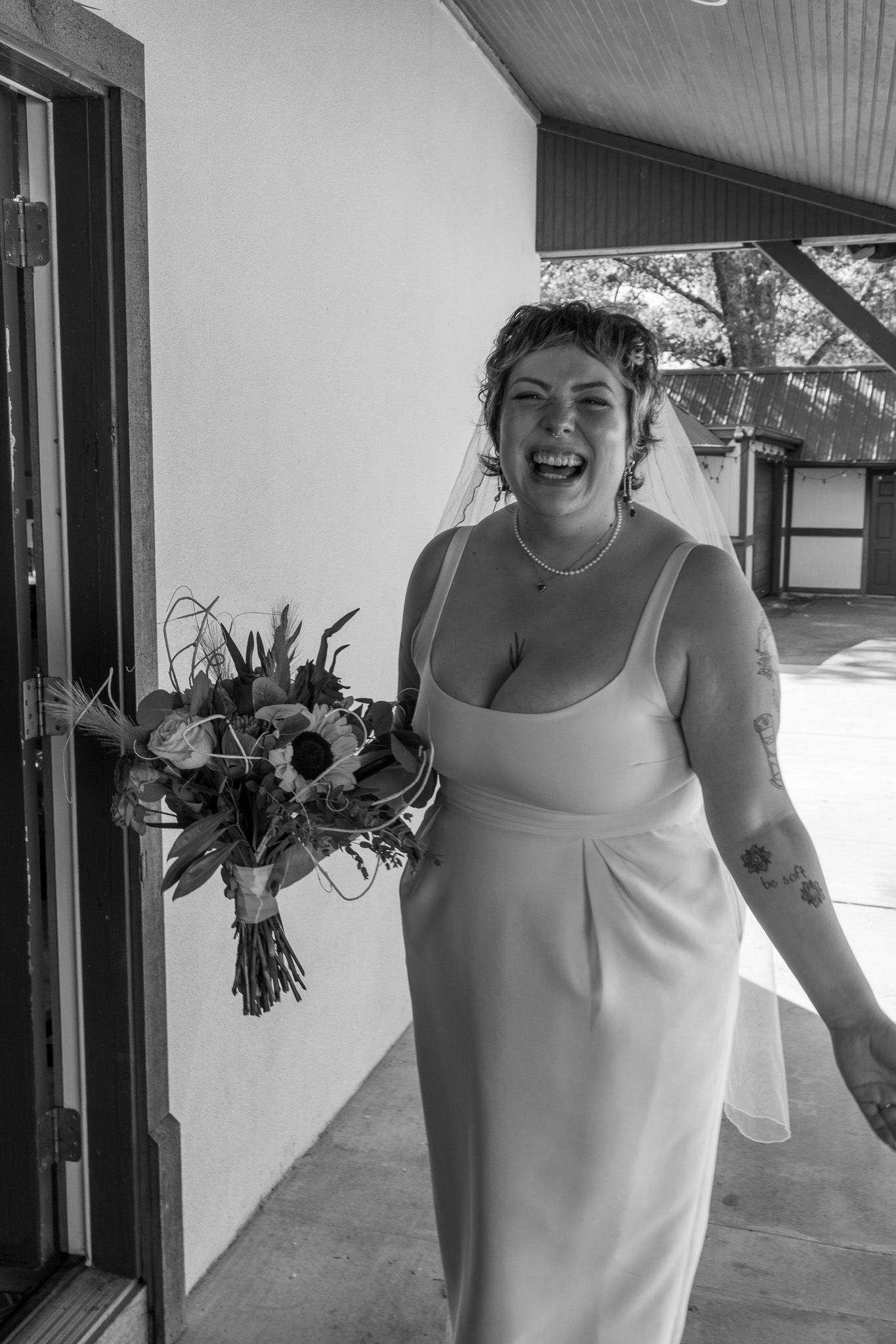 Laughing bride in white gown holds bouquet of flowers in black and white wedding portrait.