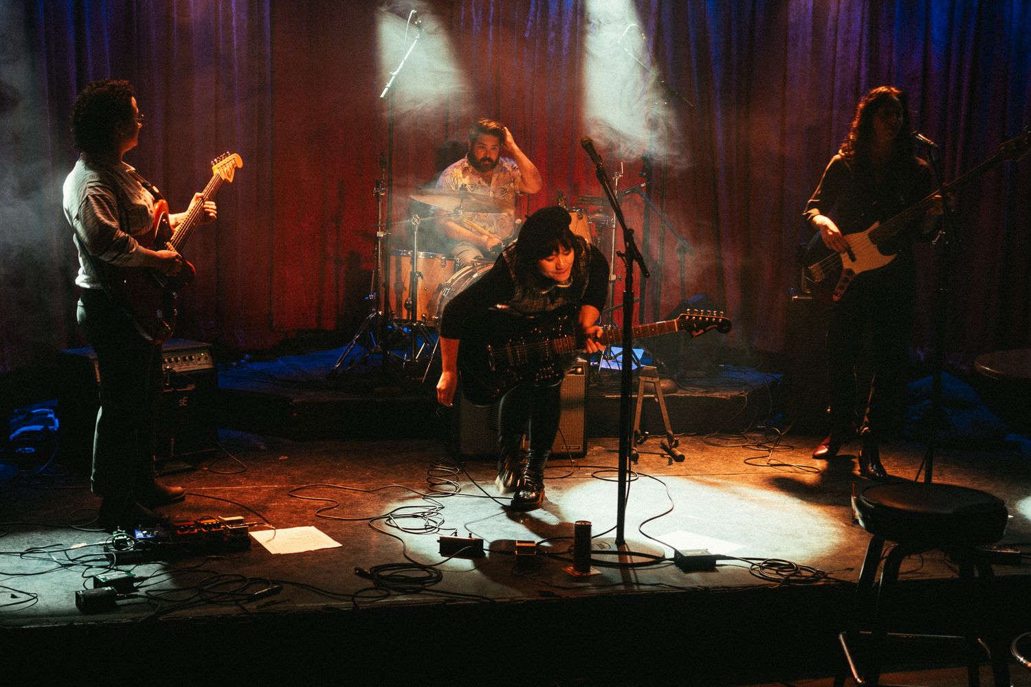 Four-piece band performing on stage with dramatic red and blue lighting and atmospheric smoke.