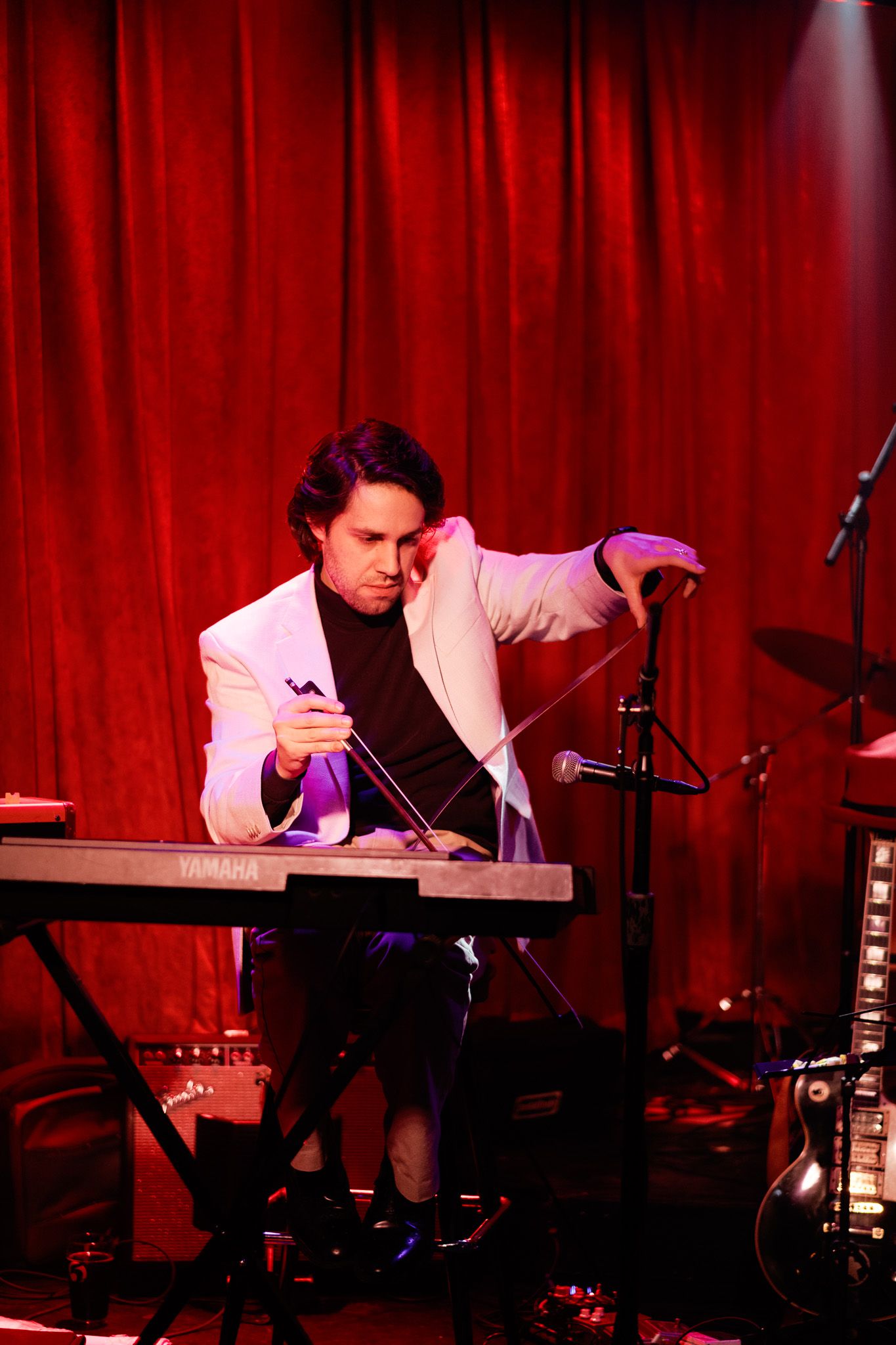 Male musician in white blazer playing a Yamaha keyboard on stage with red curtain backdrop.
