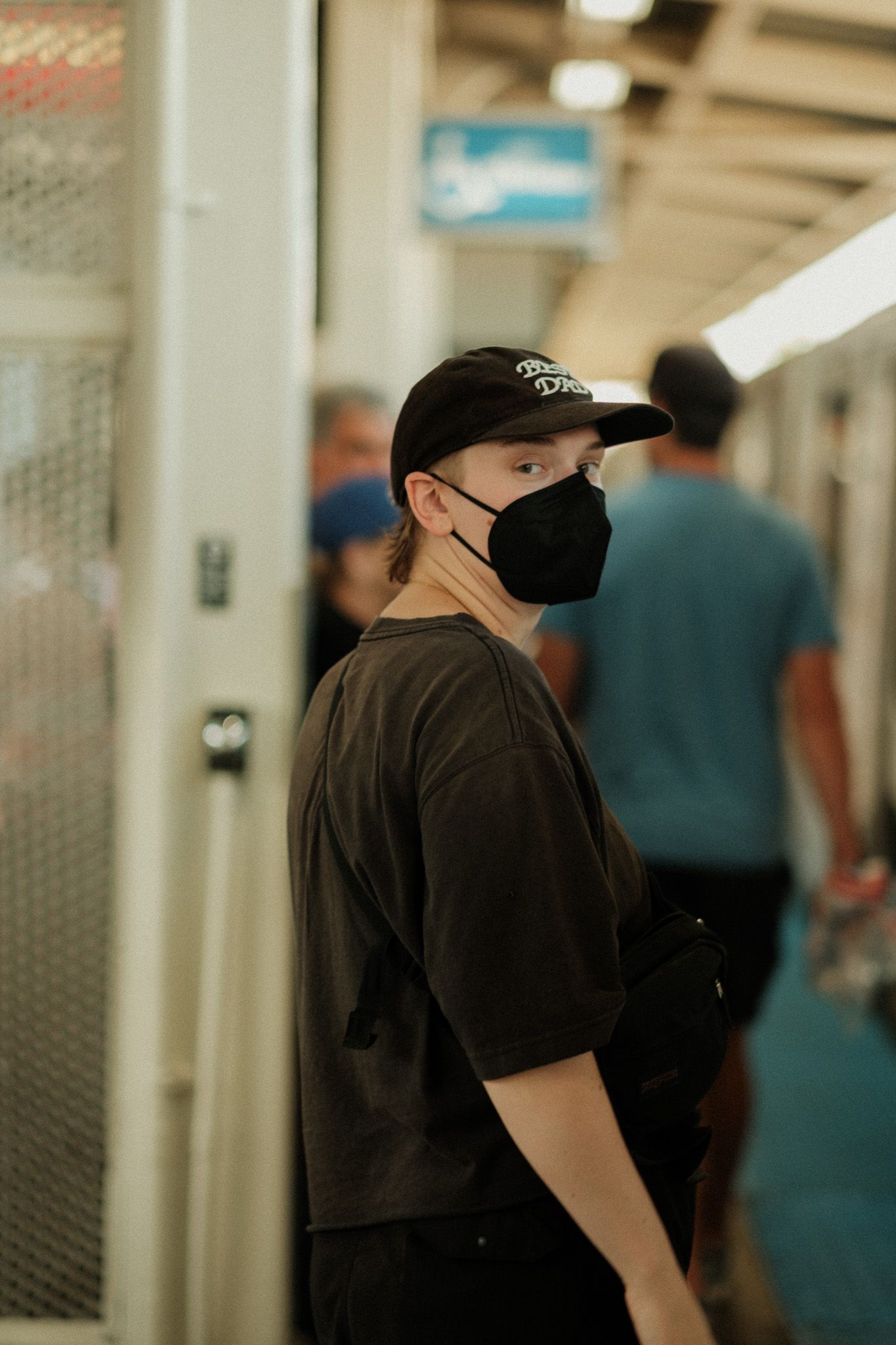 Young person wearing black mask and cap looking back over shoulder in a transit station.