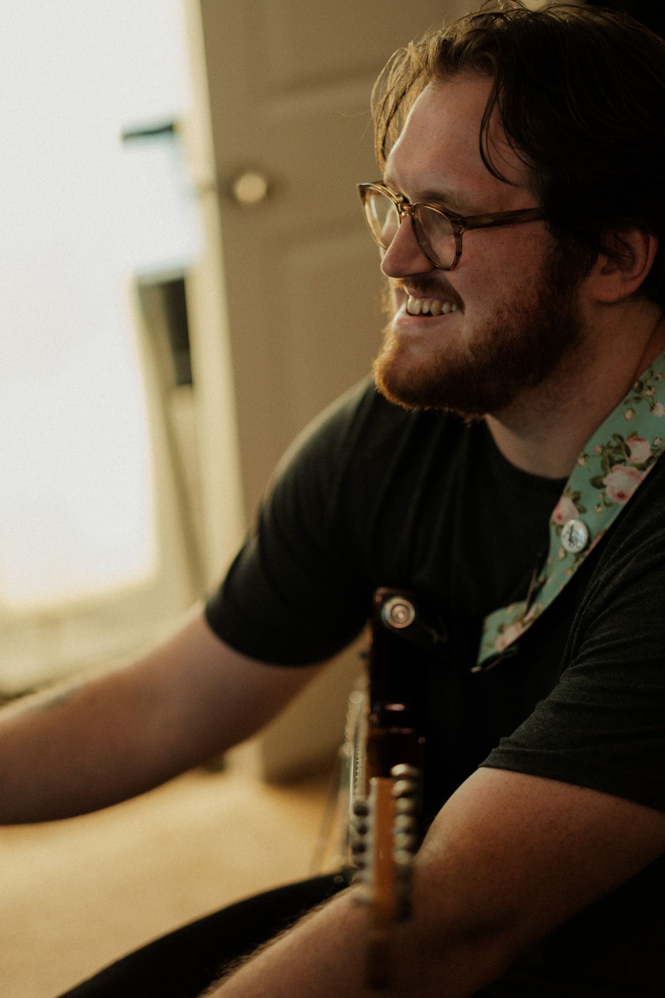 Smiling young man with glasses and beard playing guitar indoors in warm natural light.