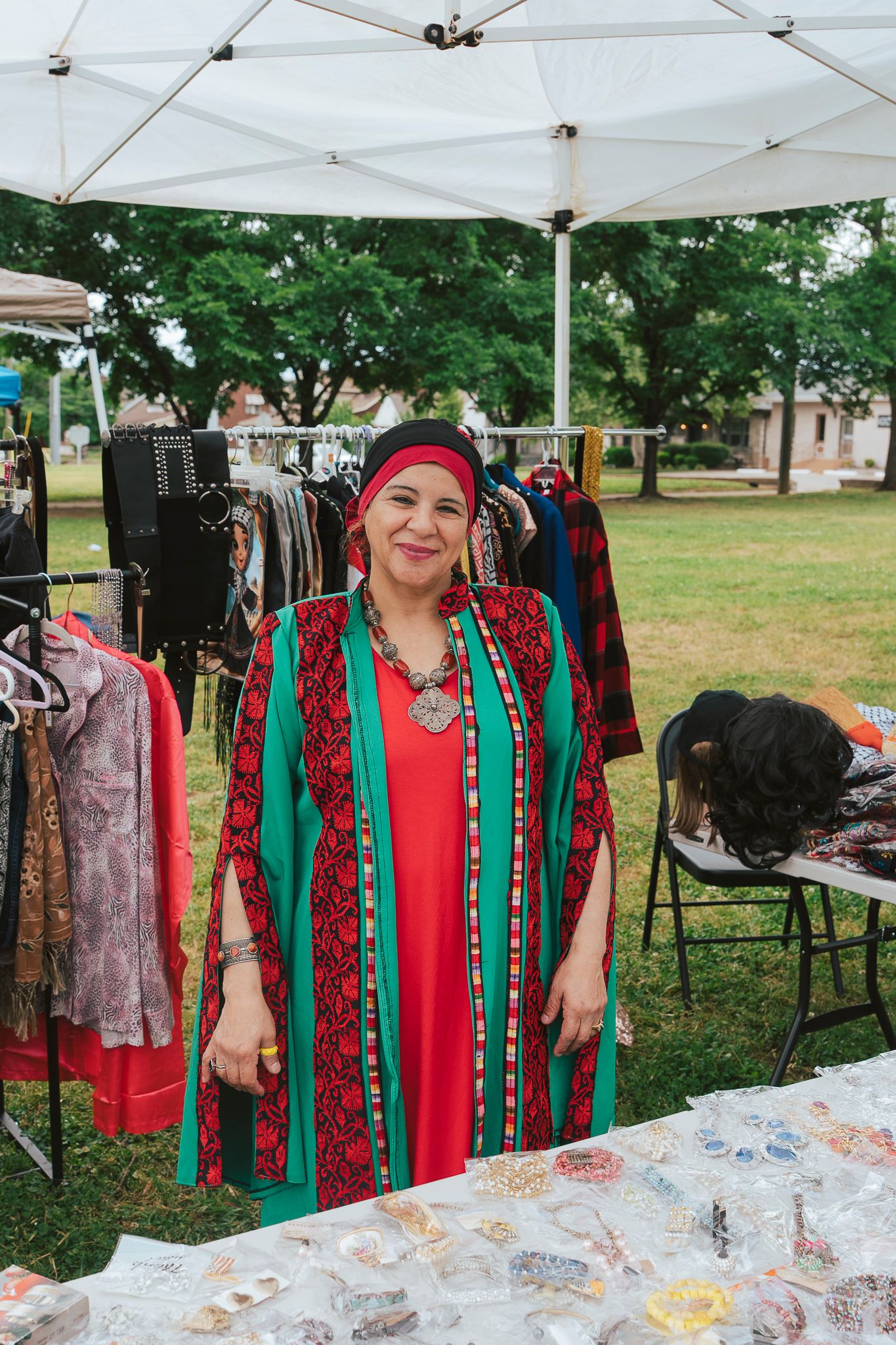 Woman in traditional red and green embroidered dress smiling at outdoor market vendor booth with jewelry and clothing.