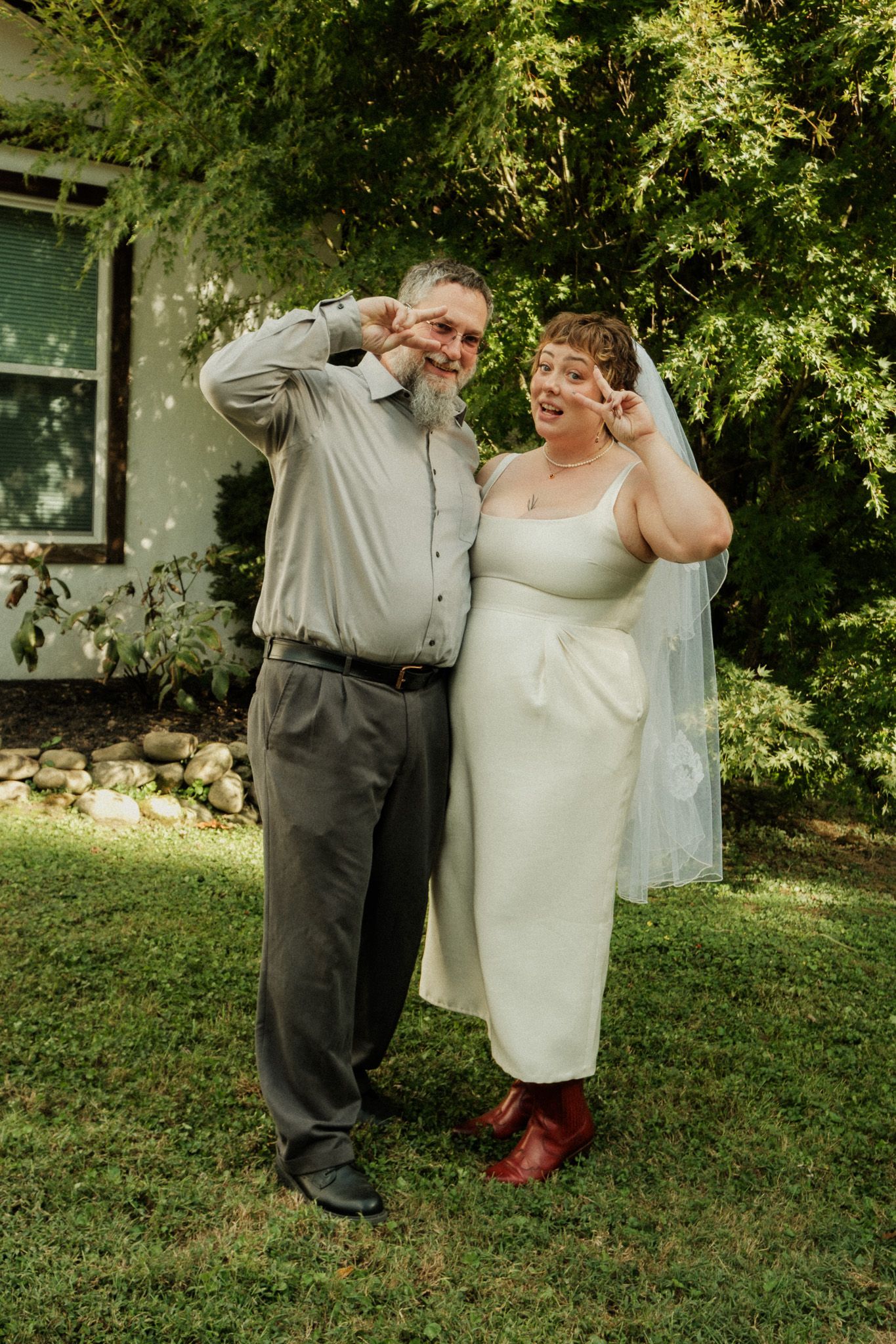 Bride in white midi dress and groom making peace signs and smiling in front of greenery.