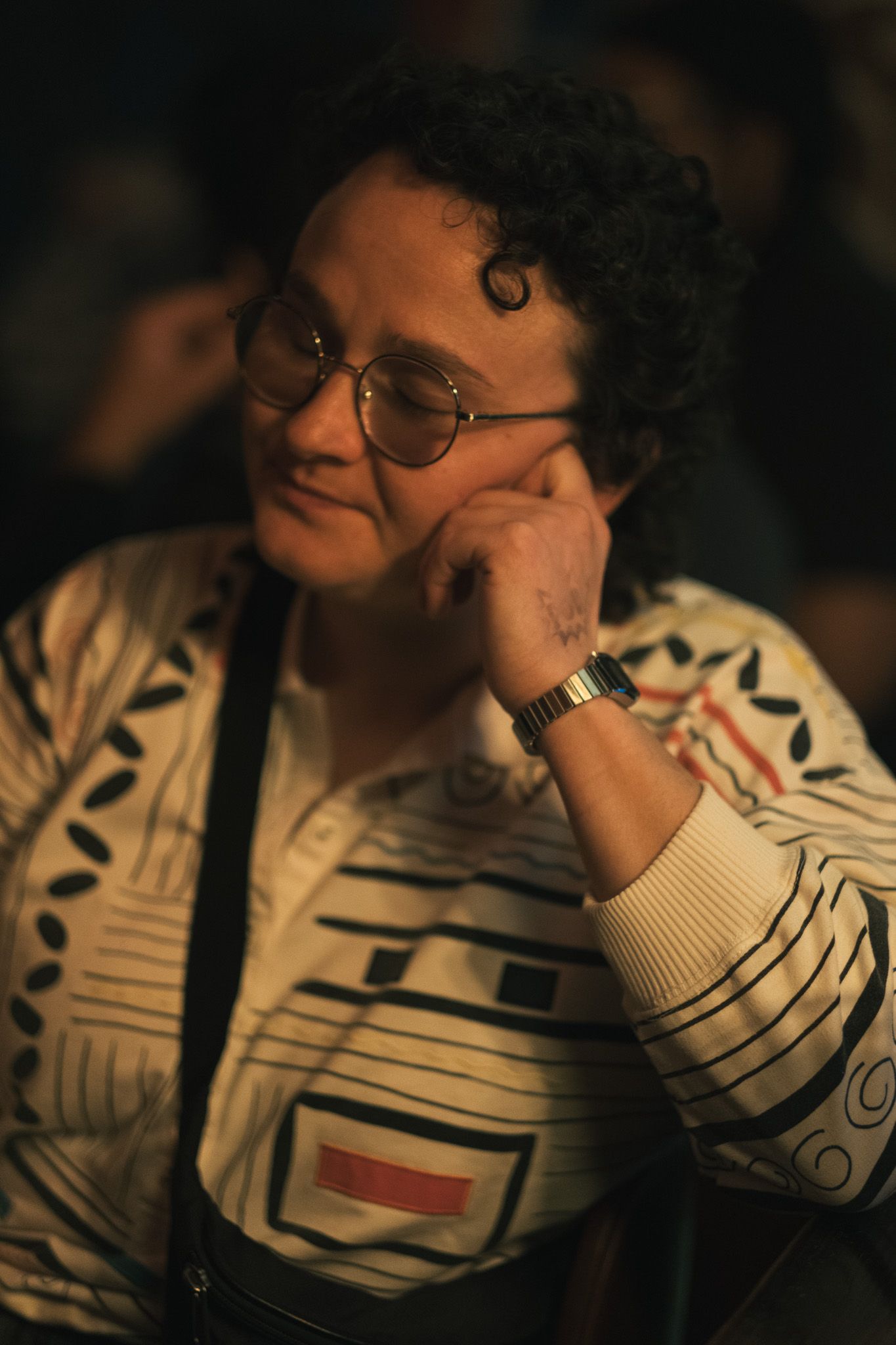 A person with curly hair and round glasses rests their chin on their hand thoughtfully in a dimly lit room.