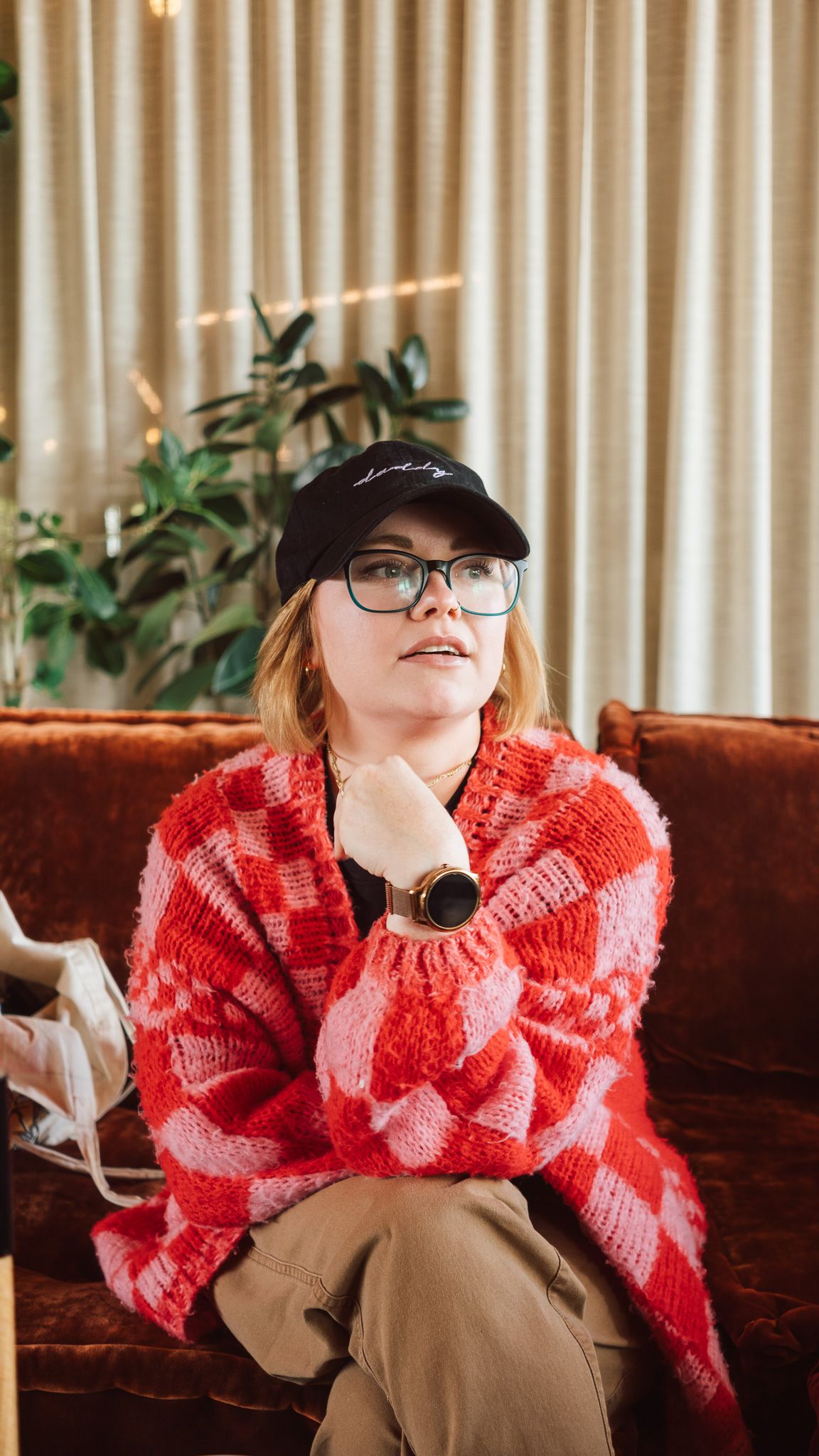 Woman in a red and pink checkered knit sweater and black cap sitting on a couch, wearing glasses and a watch.