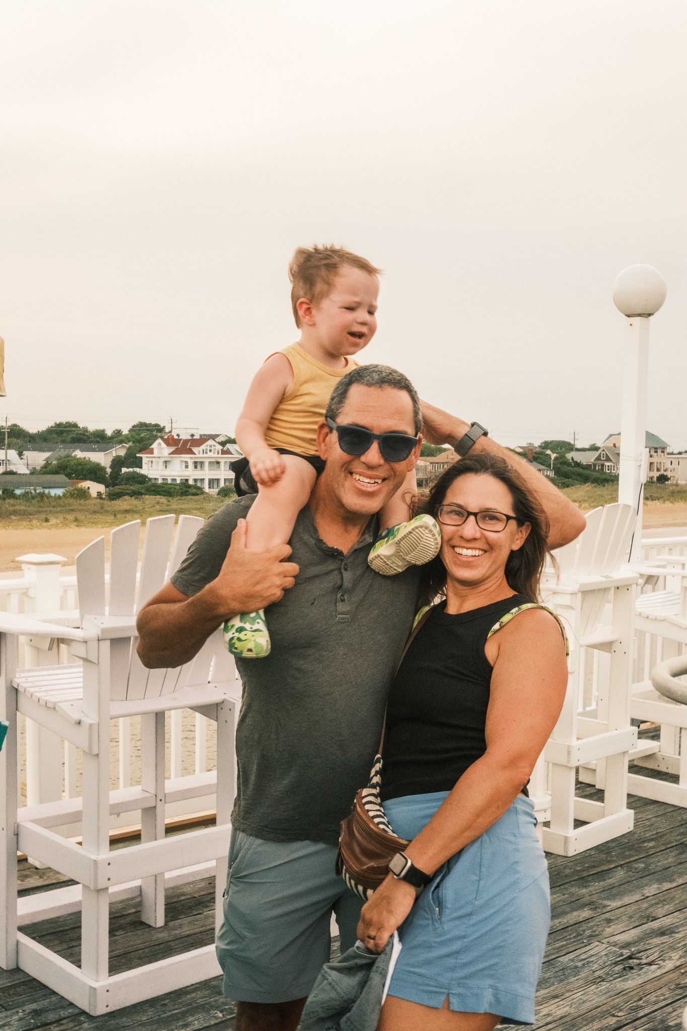 Happy family with toddler on dad's shoulders posing on a beach boardwalk.