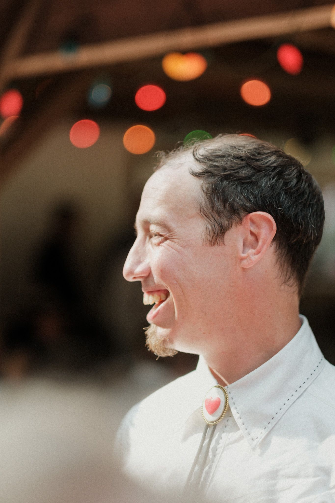 Smiling groom in white western shirt with heart bolo tie laughing joyfully at wedding reception.