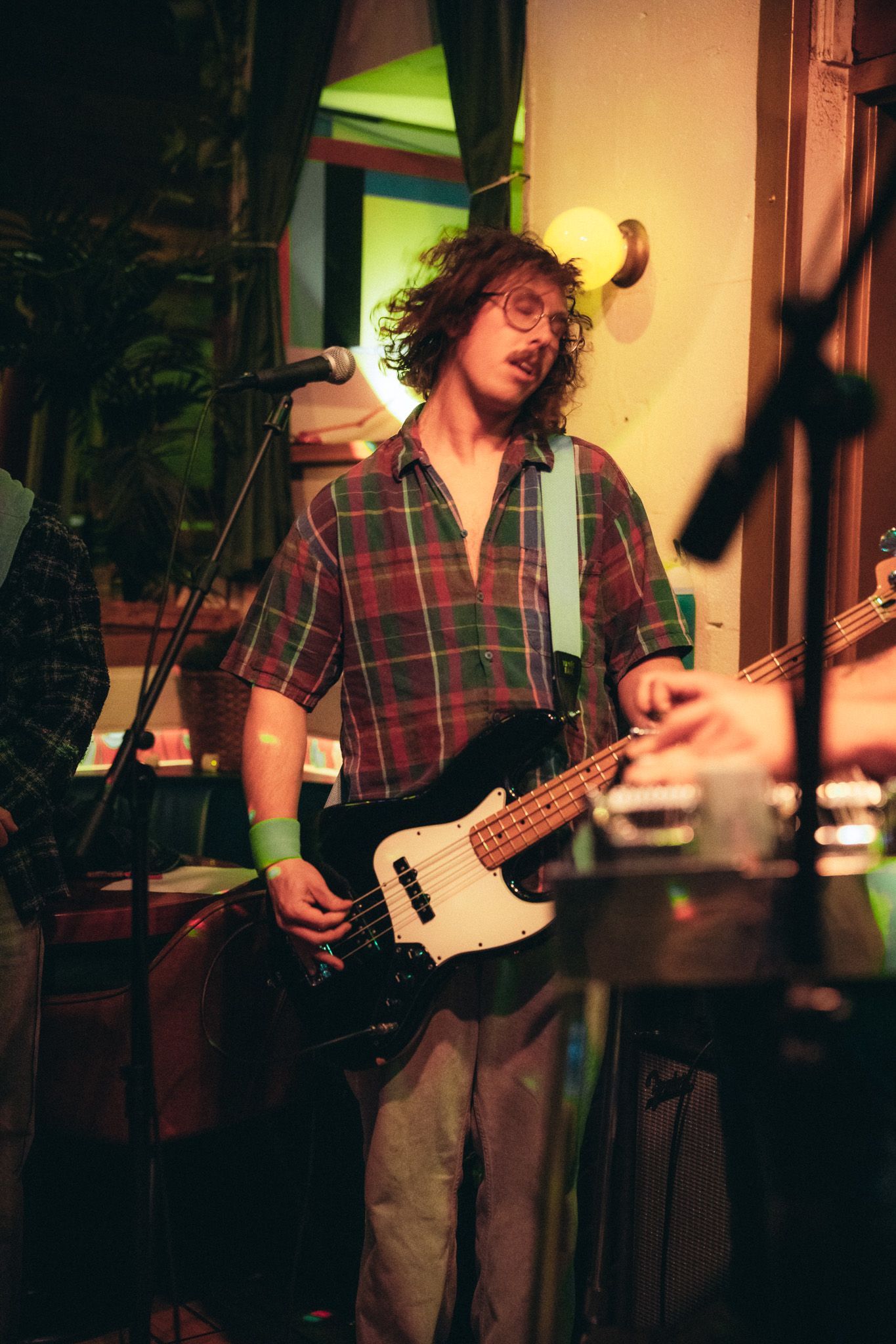 Curly-haired bassist in plaid shirt playing electric bass guitar at a cozy indoor venue.