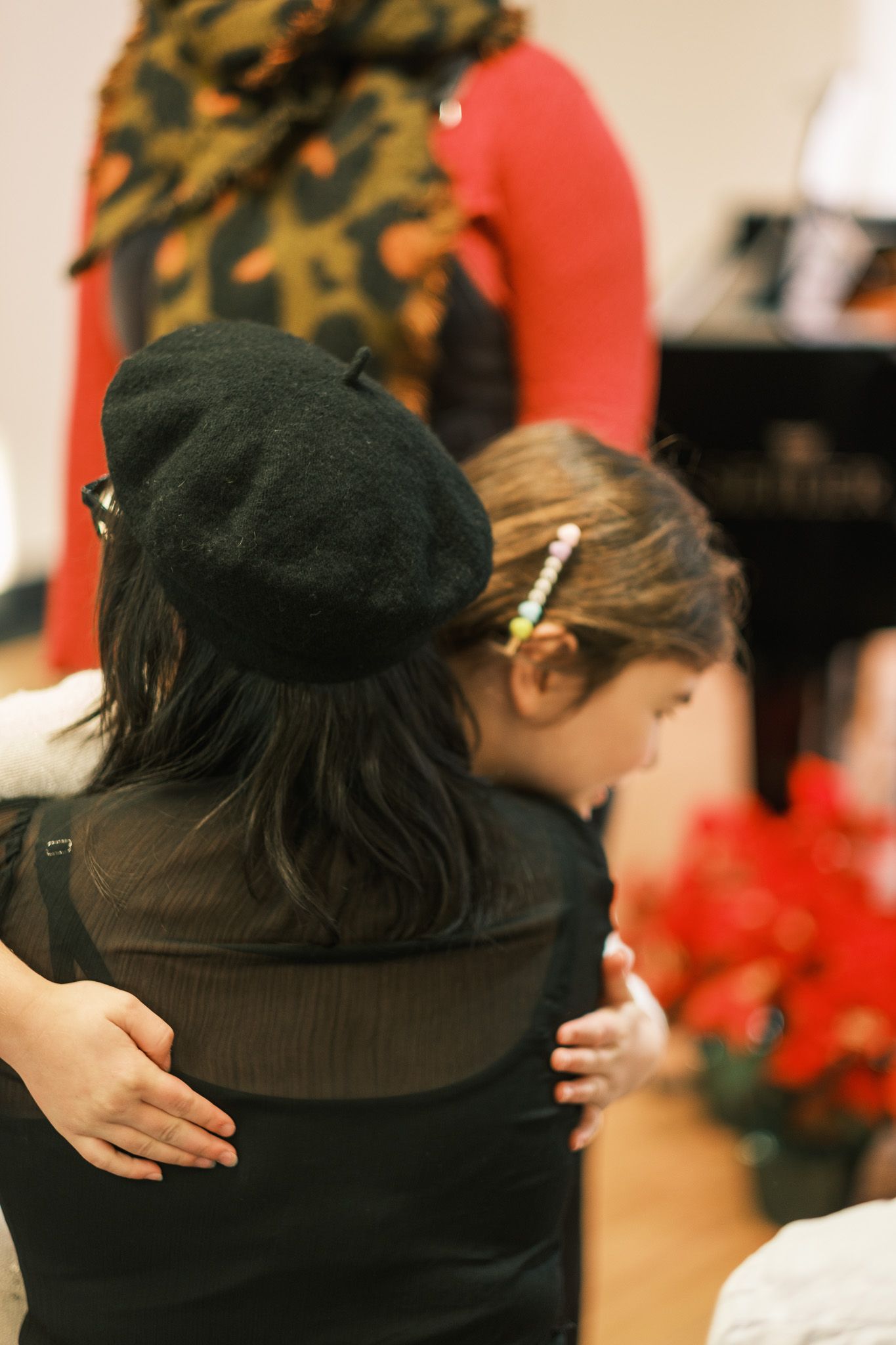 Woman in black beret embraces a young girl with hair clips at a holiday music recital near red poinsettias.