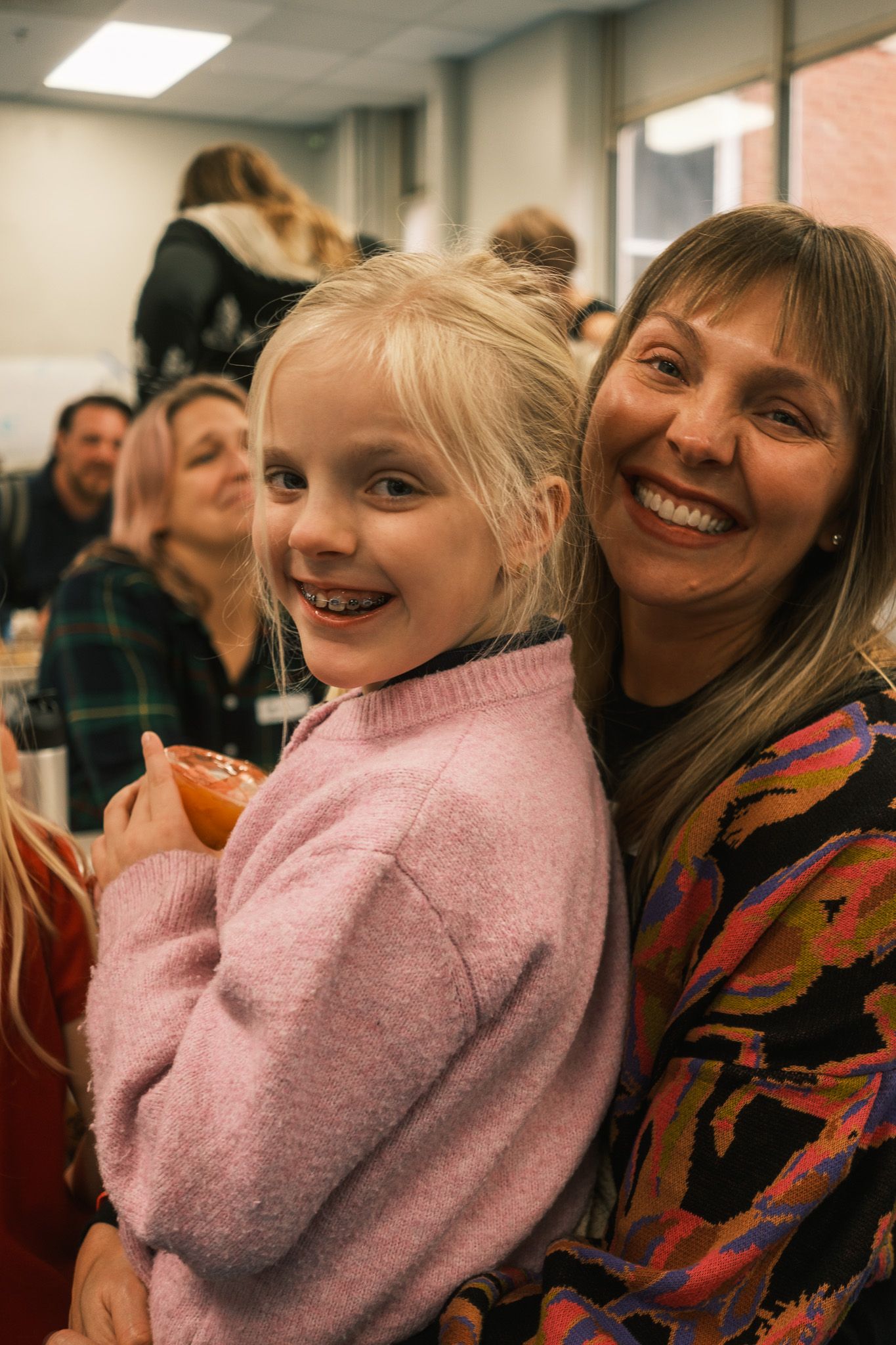 Smiling woman hugging a young blonde girl in a pink sweater at an indoor event.