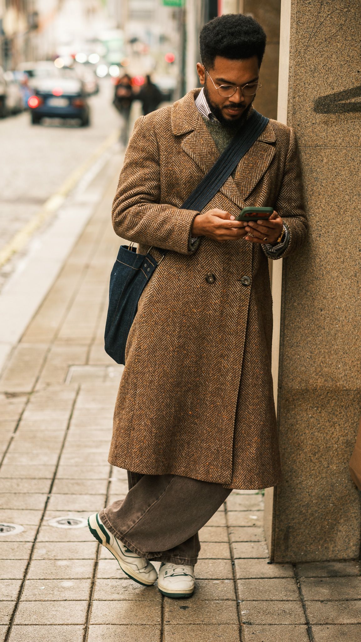 Stylish man in a long brown tweed coat using his phone while leaning against a city wall.