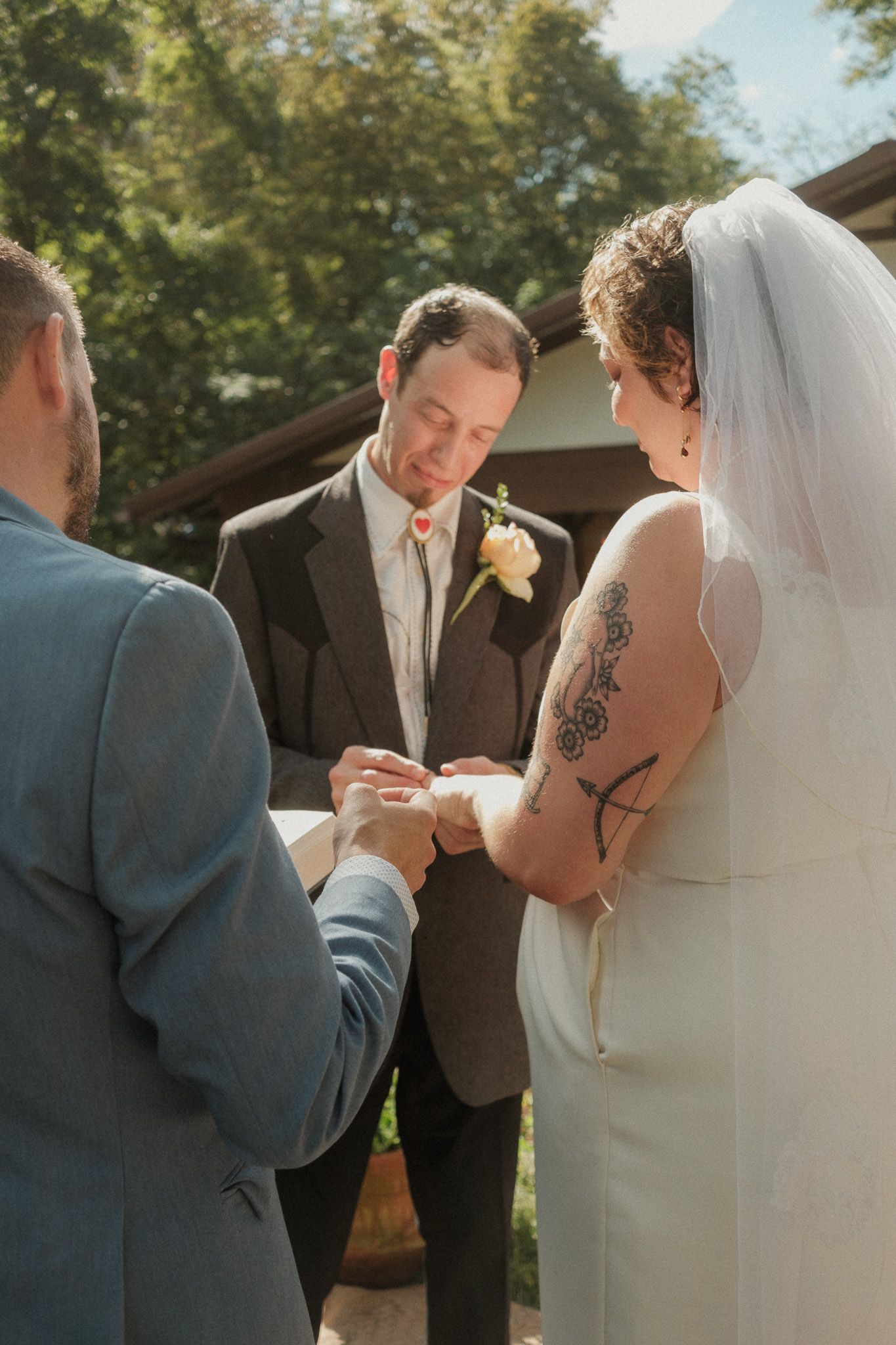 Groom places wedding ring on brides finger during outdoor ceremony while officiant watches closely.