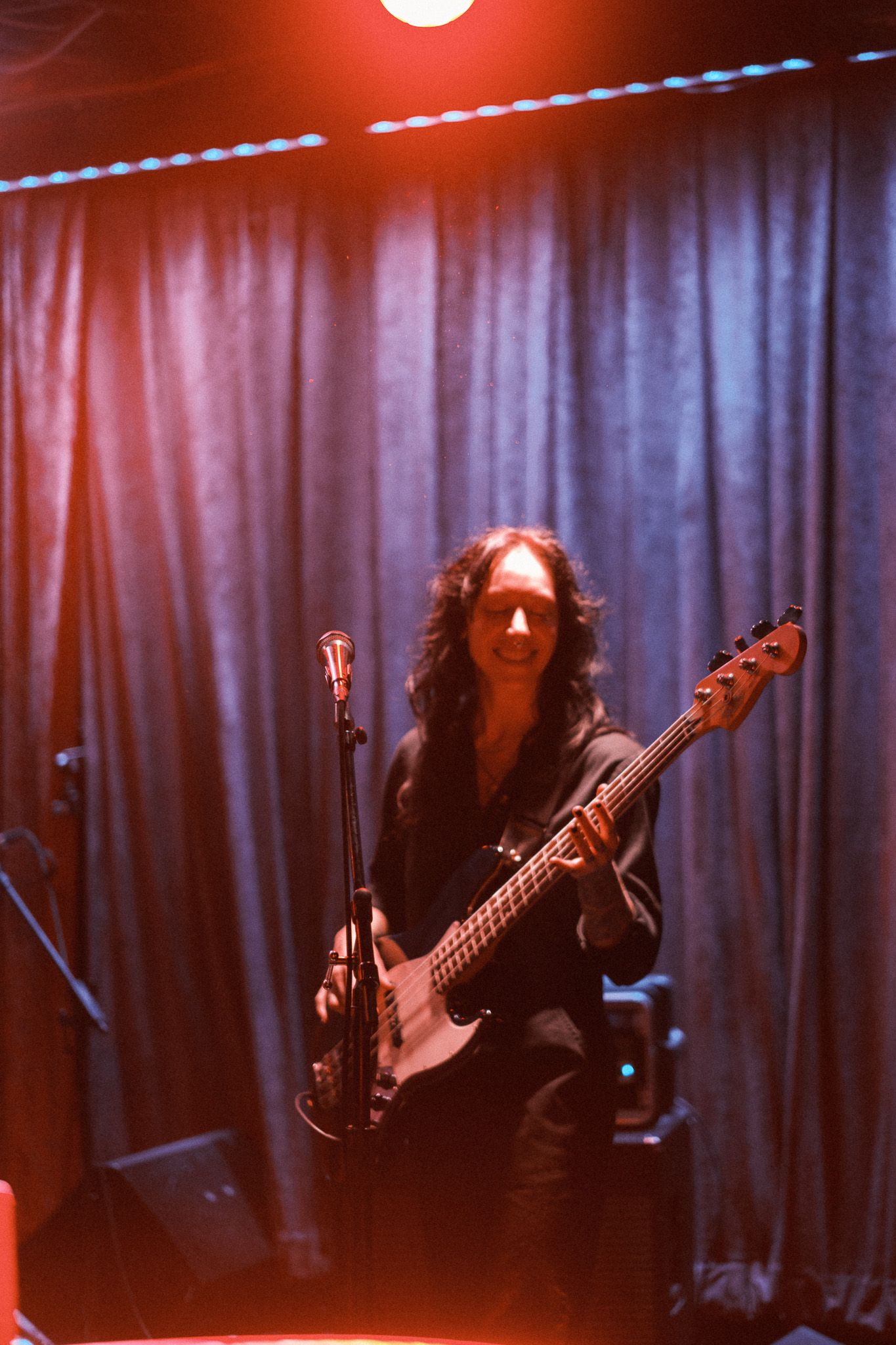 Female bassist in black outfit performing on stage under red lighting with blue curtain backdrop.