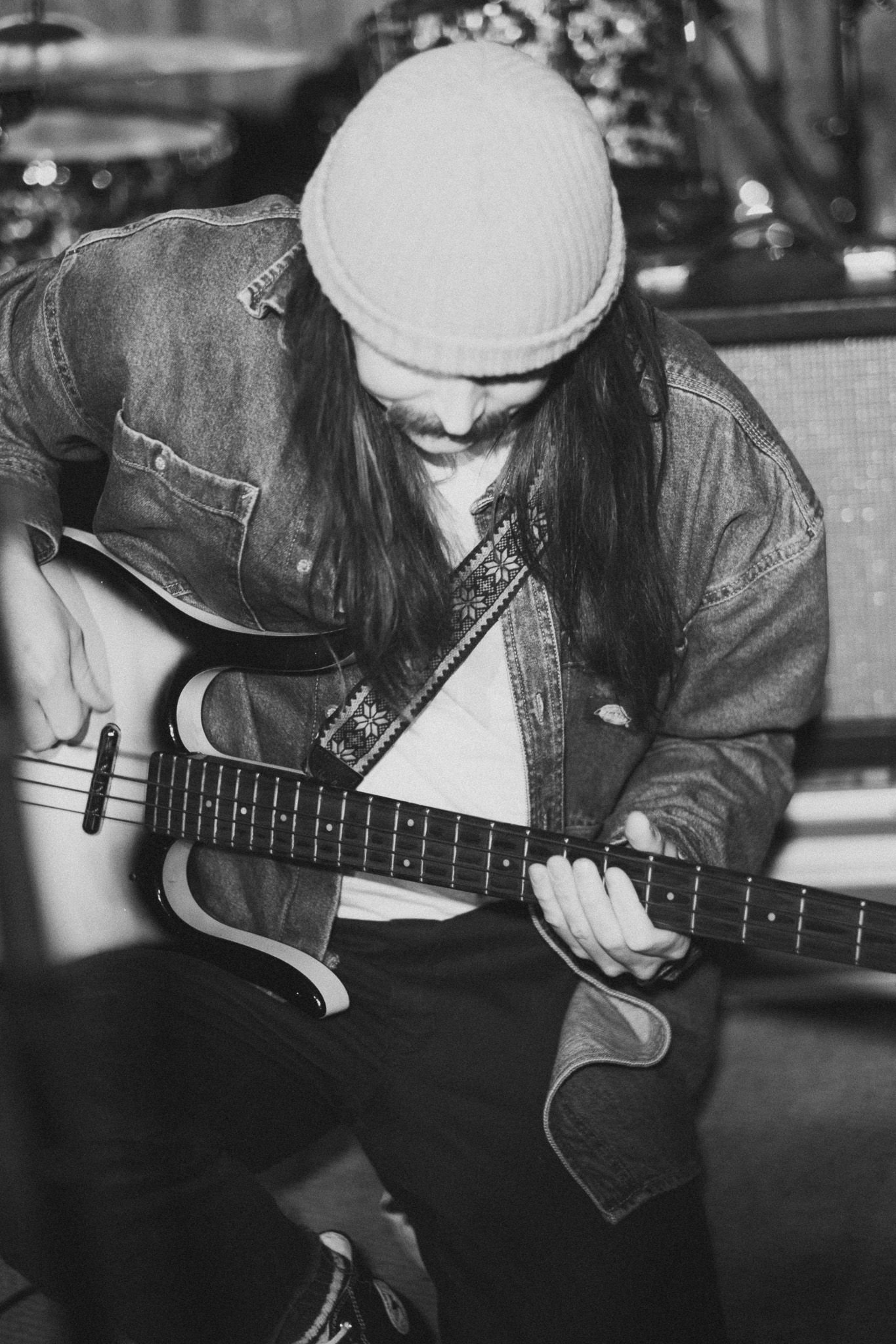 Long-haired guitarist in denim jacket and beanie focuses on playing bass in black and white photo.