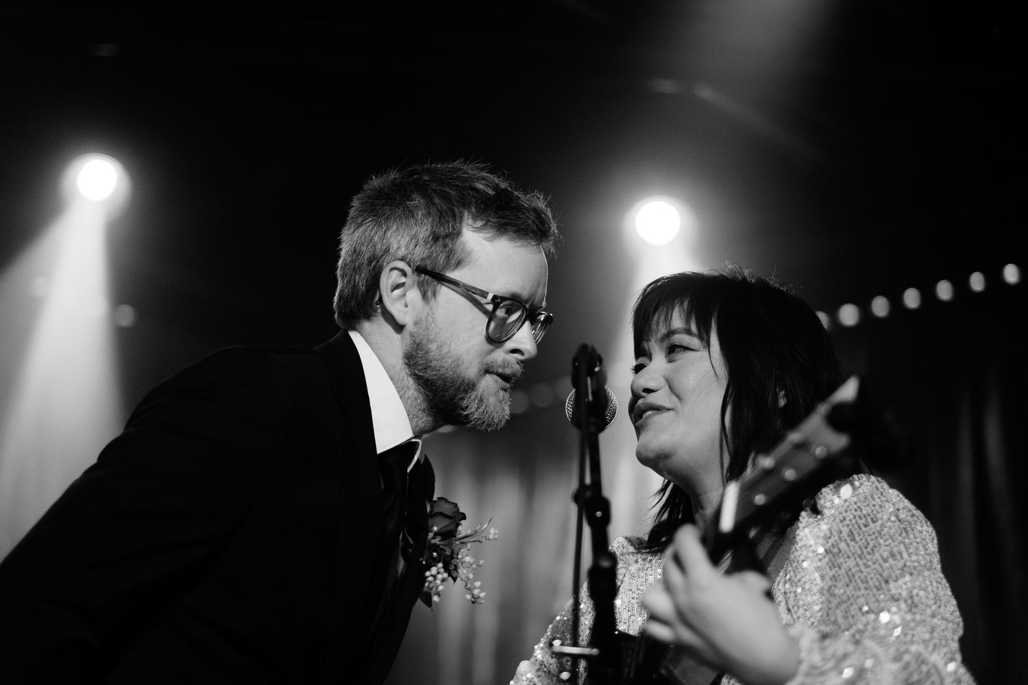 Black and white photo of two musicians sharing a microphone on stage under spotlights.