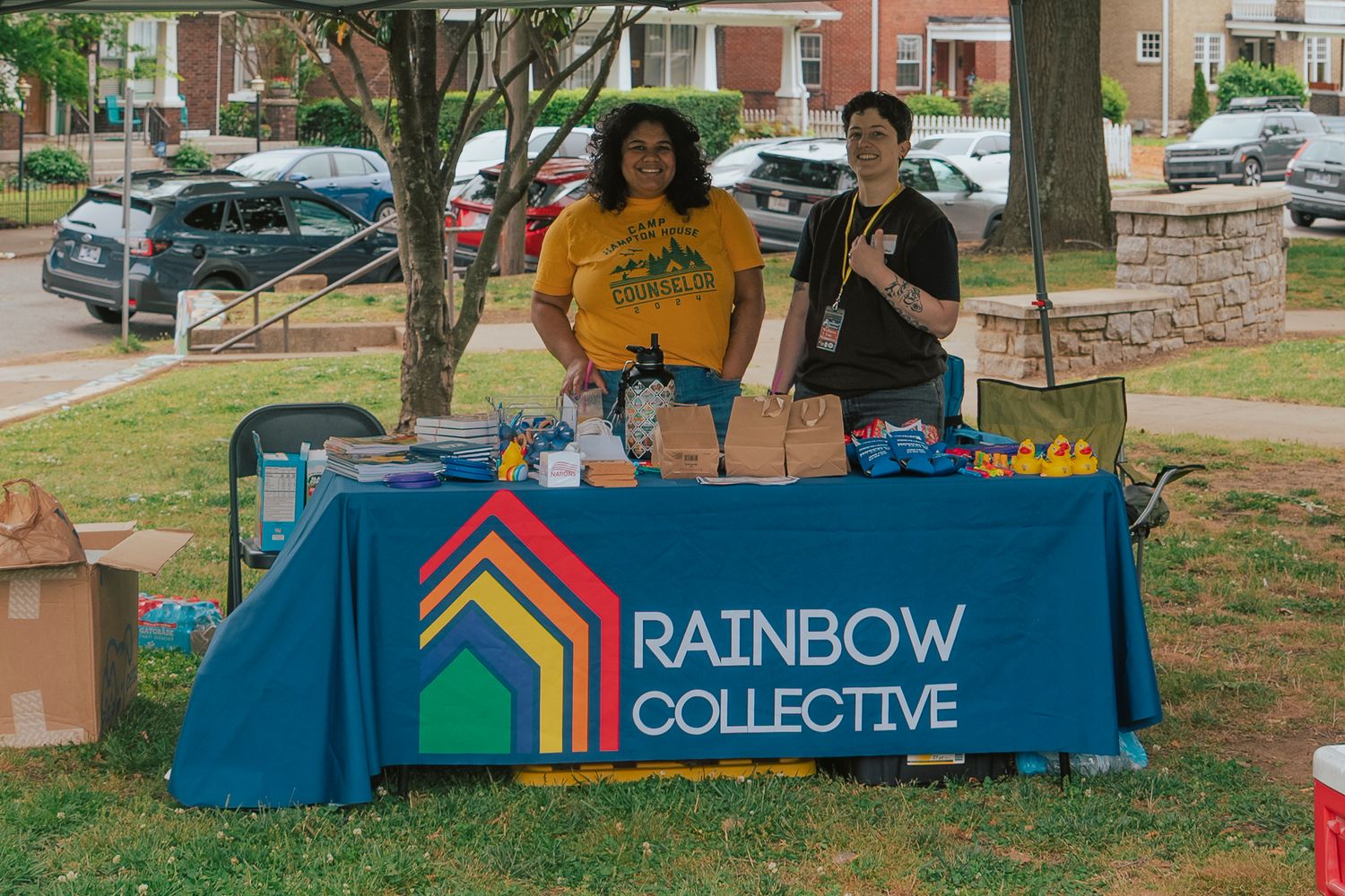 Two women in yellow shirts at Rainbow Collective booth with colorful logo at outdoor event.