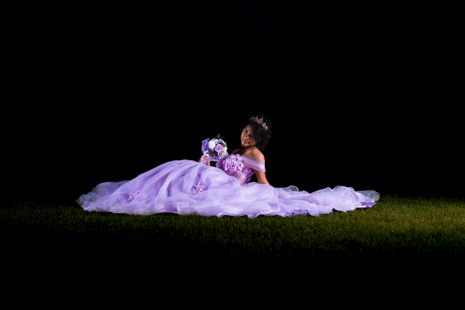Teen in flowing lavender ball gown sits gracefully on grass under dramatic lighting against a dark background in McKinney, TX by Kin & Lens