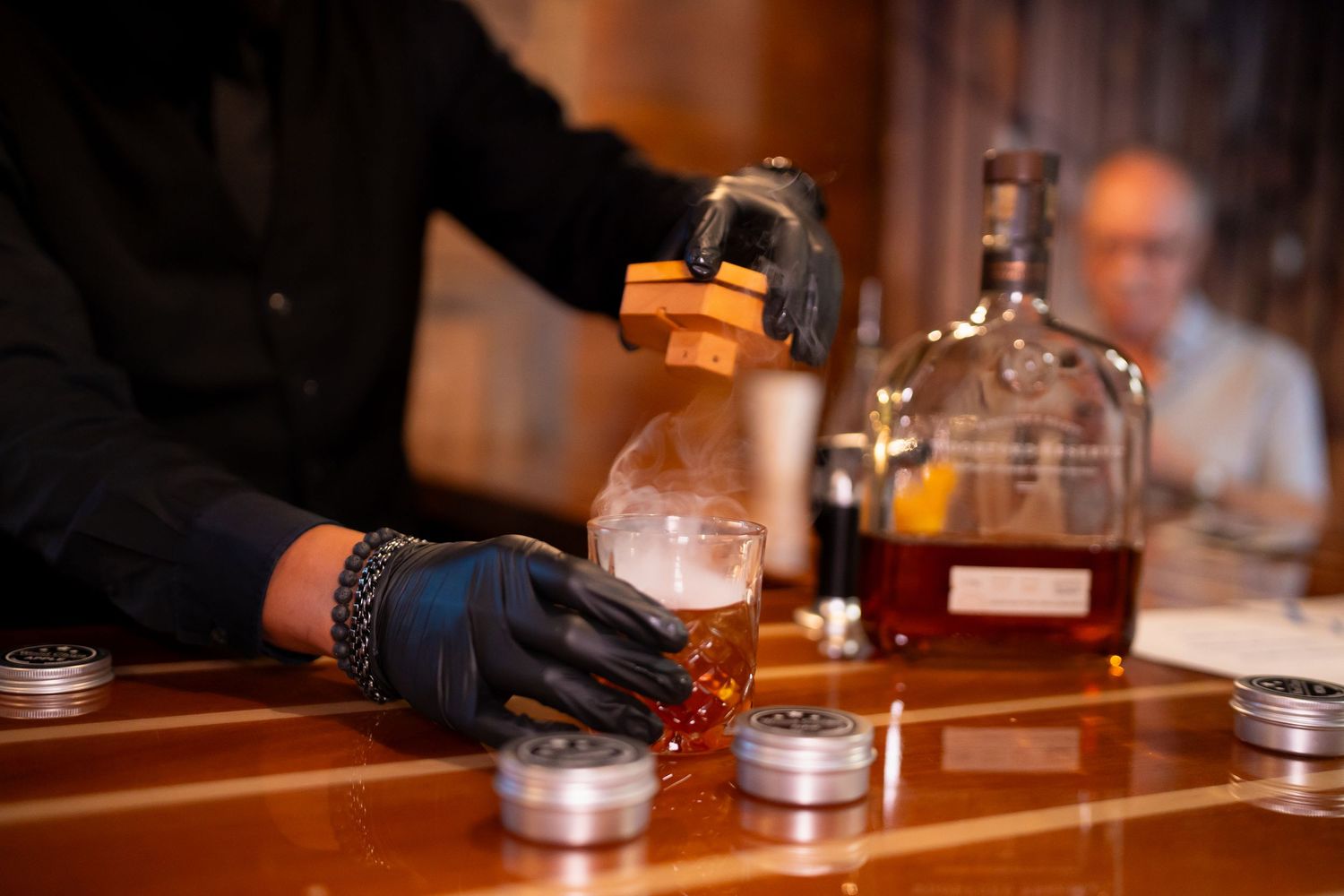 Bartender wearing black gloves pours a cocktail in a glass at a wooden bar with bottles in the background in Addison by Kin & Lens Creative