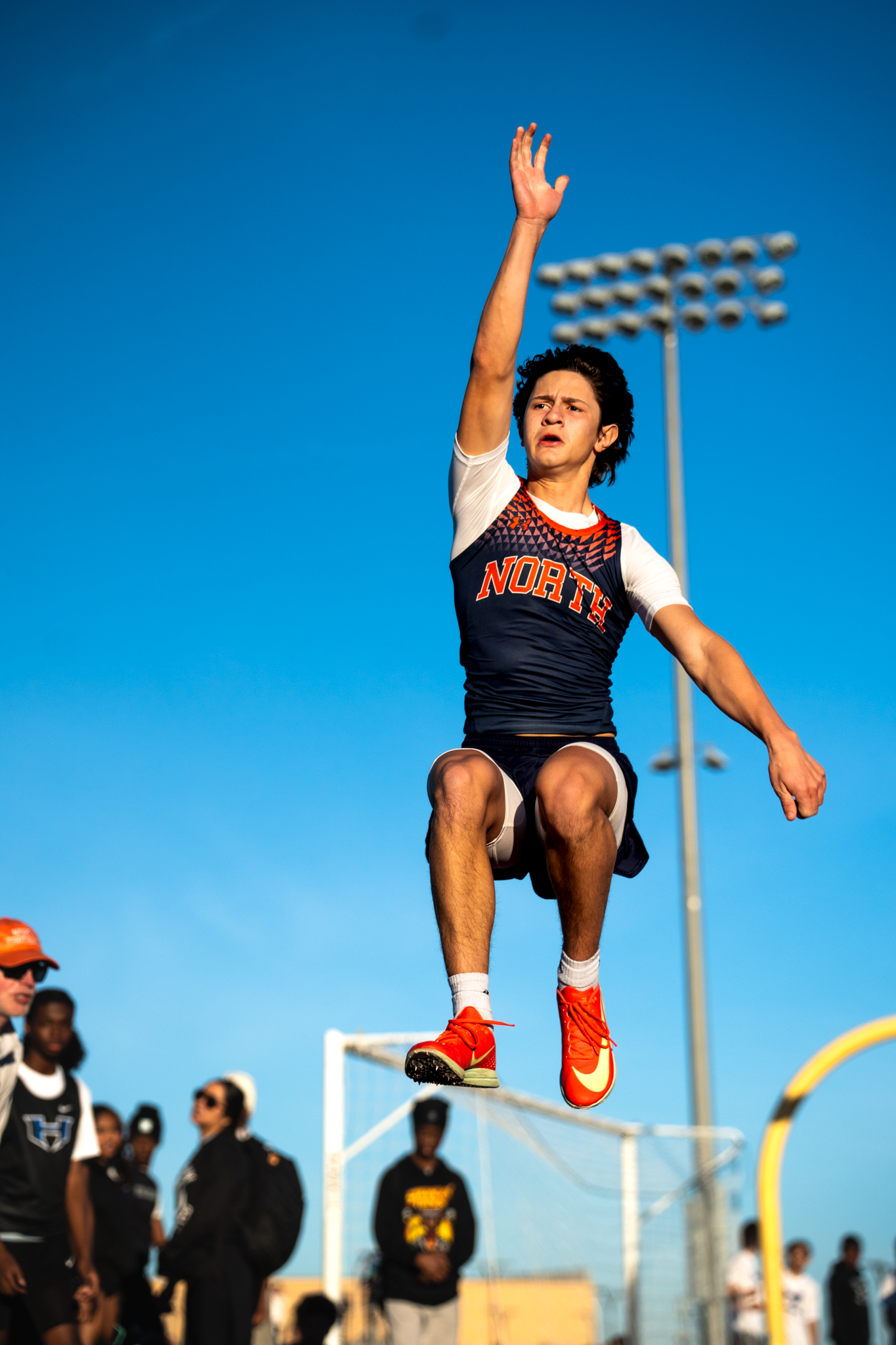 Athlete in red and black uniform performs dynamic long jump against bright blue sky and stadium lights McKinney by Kin & Lens Creative Media