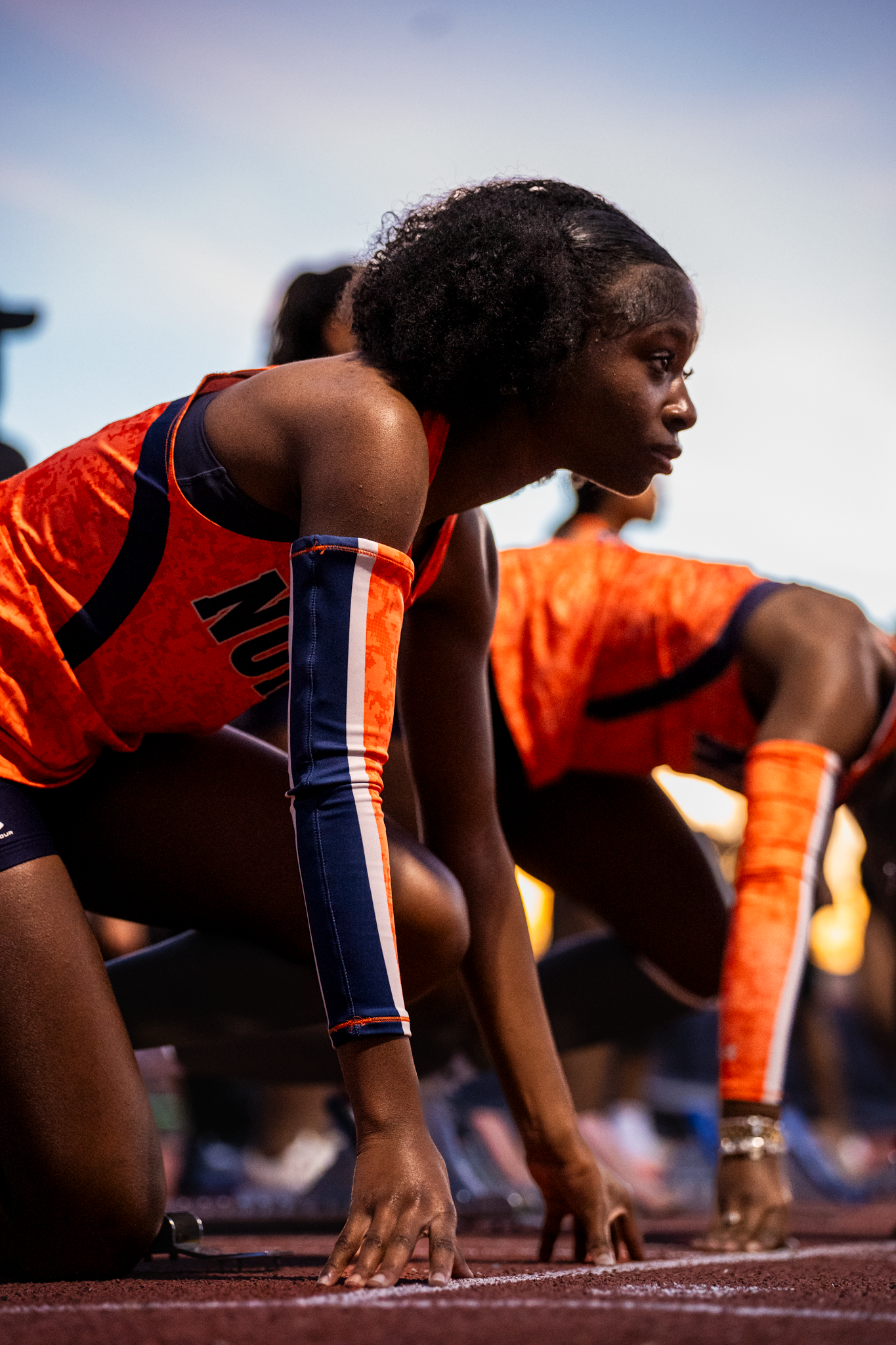 Athletes in orange uniforms line up at starting position on a track.