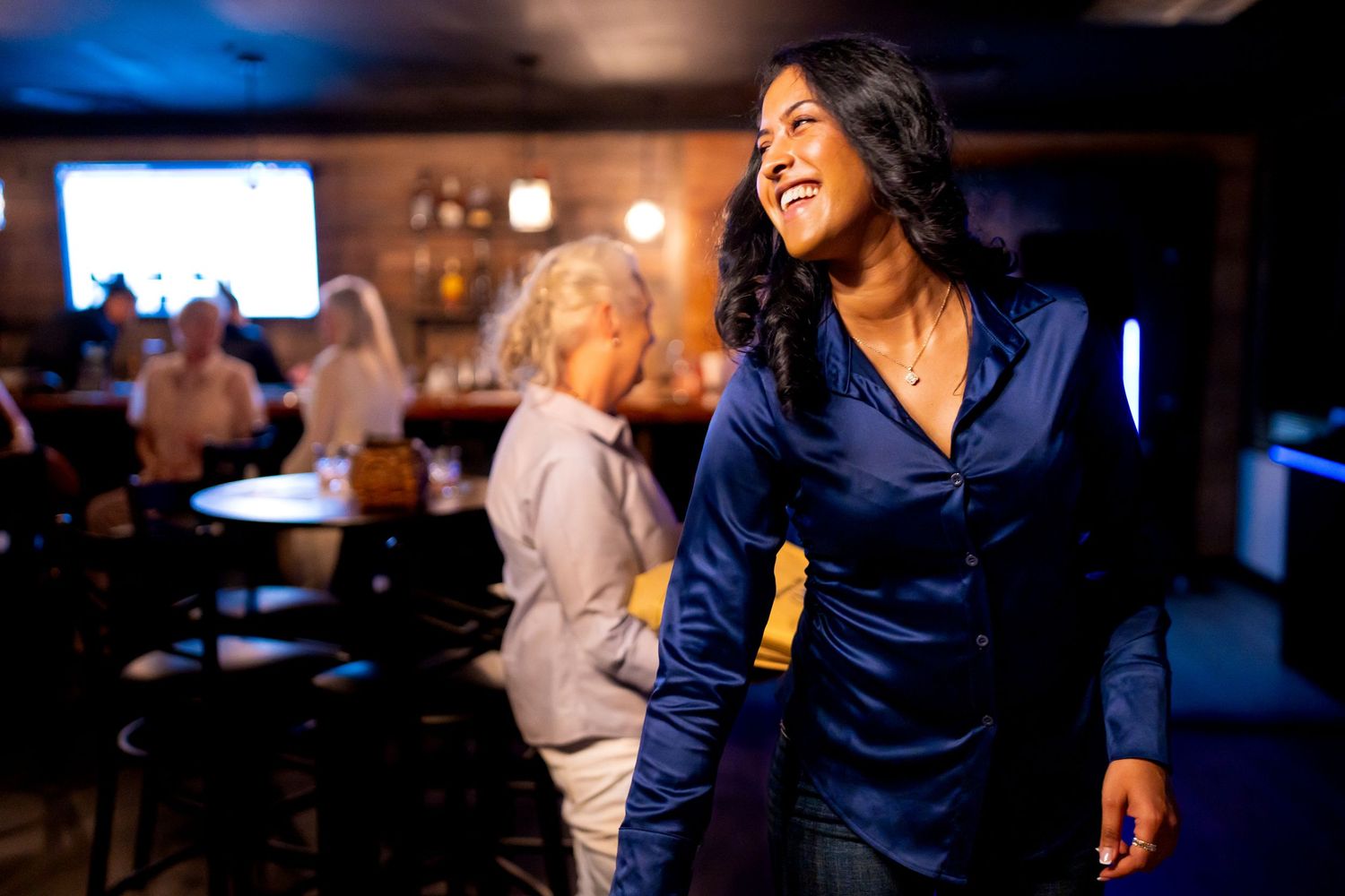 Smiling woman in blue blouse enjoying a lively social gathering in a dimly lit venue in Addison, TX by Kin & Lens Creative Media