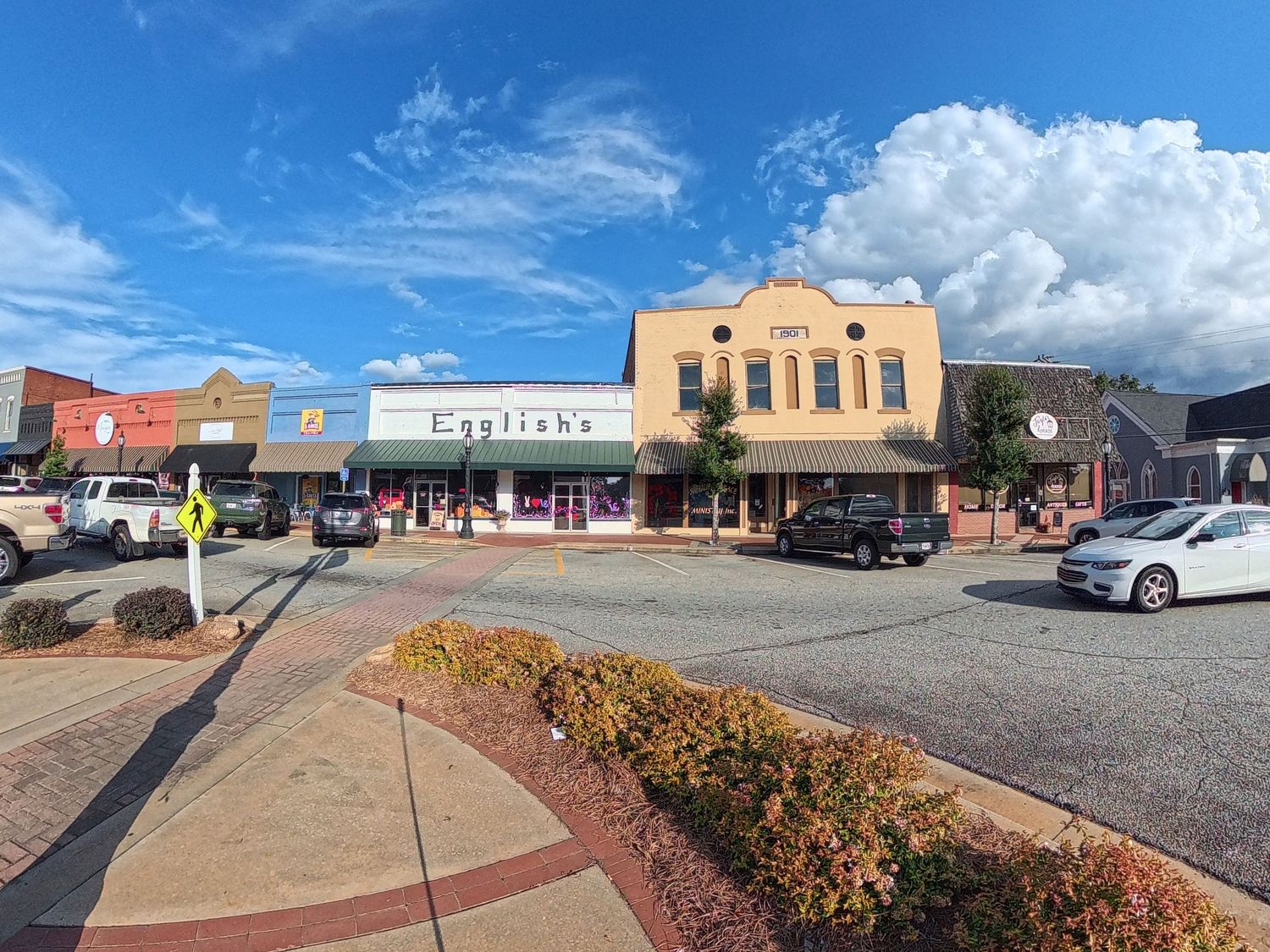 Panoramic view of a small town main street with colorful storefronts, parked cars, and yellow flowering bushes along sidewalks.