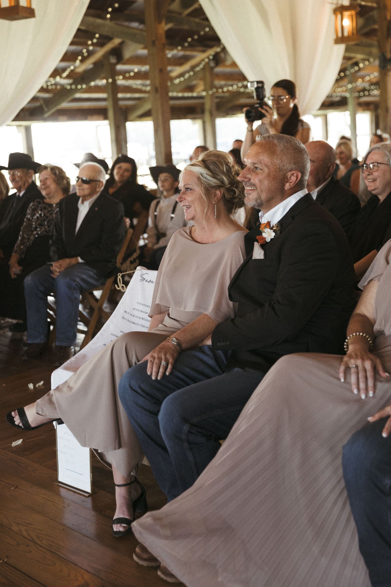 Wedding guests laugh and smile while seated in rows at a rustic barn venue decorated with white fabric draping.