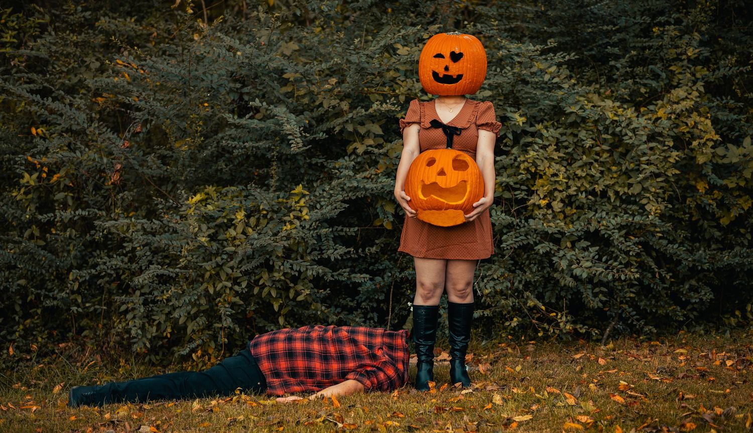 A figure in a brown dress holds a jack-o'-lantern bucket while standing in autumn foliage with a plaid blanket nearby.