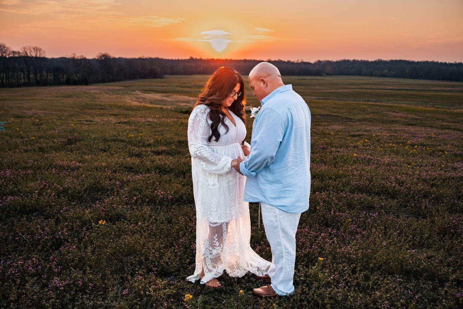 Couple in white and blue outfits holding hands in a field at golden sunset, maternity photo session outdoors.