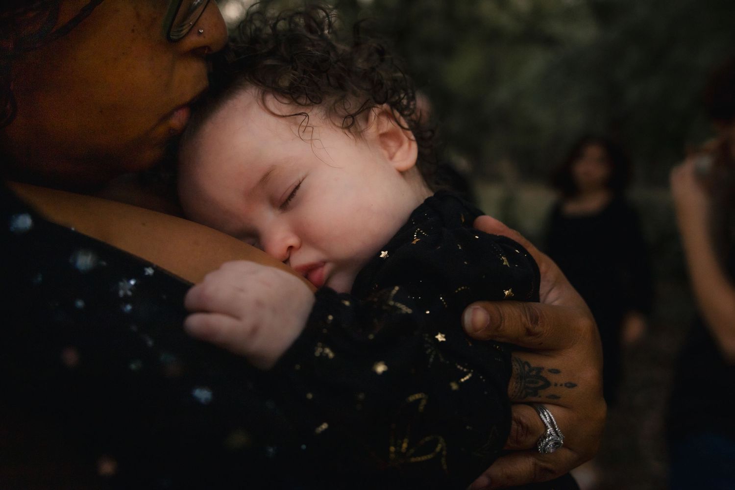 A sleeping baby resting peacefully in a parent's tattooed arms at an outdoor gathering.