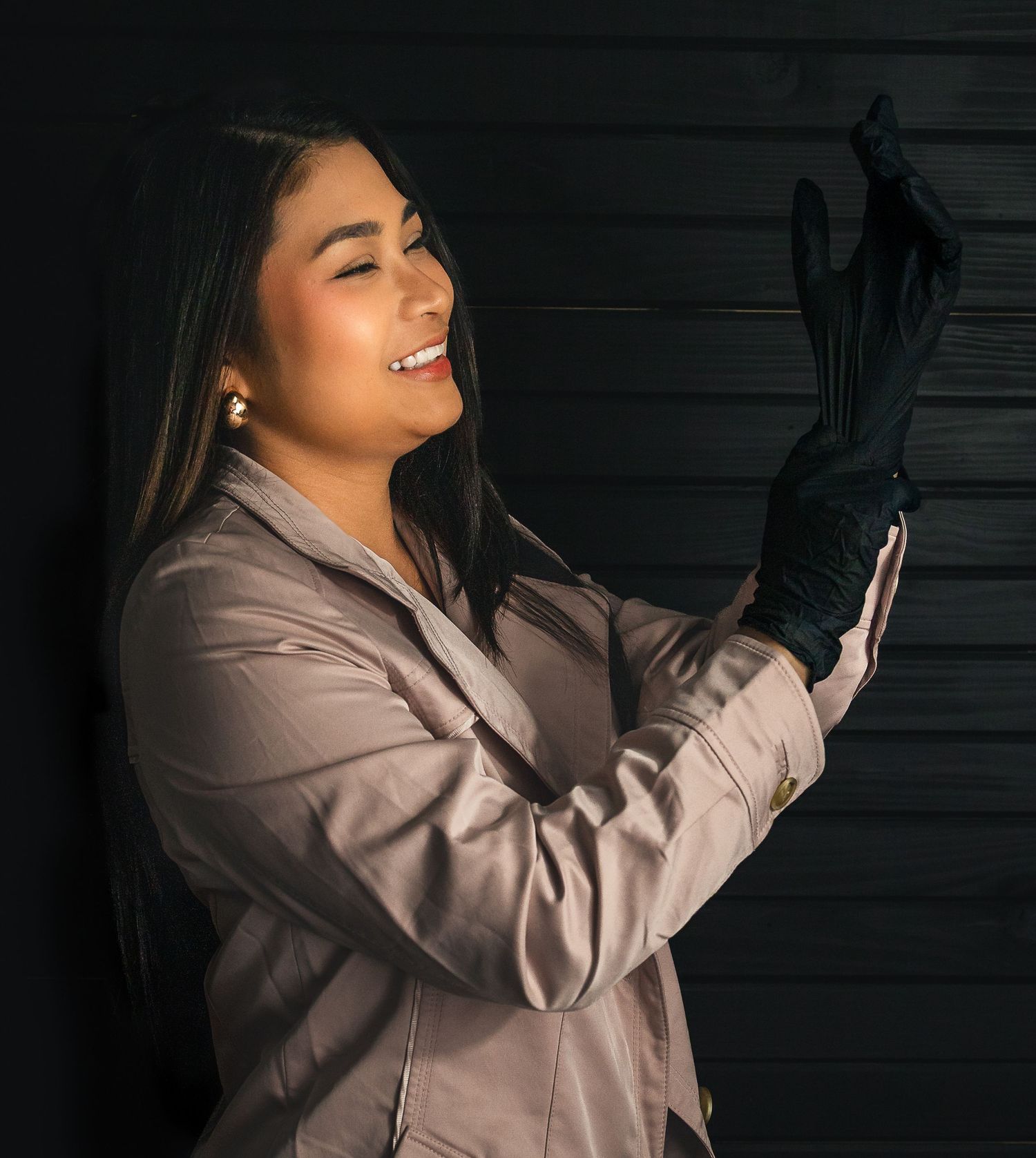 Woman in satin jacket smiling while putting on black gloves against dark background.
