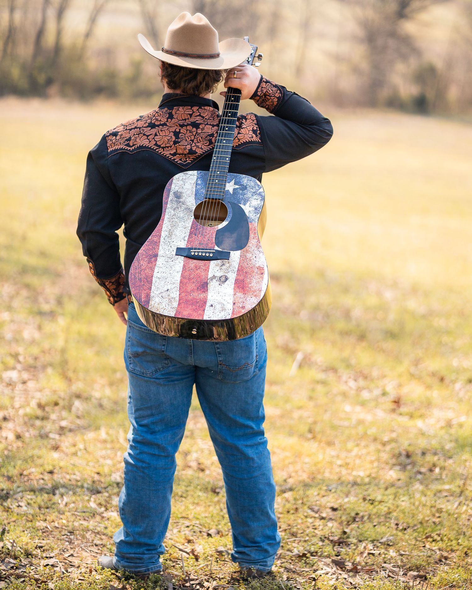 Man in cowboy hat holding American flag acoustic guitar in a field, wearing black western shirt and jeans.