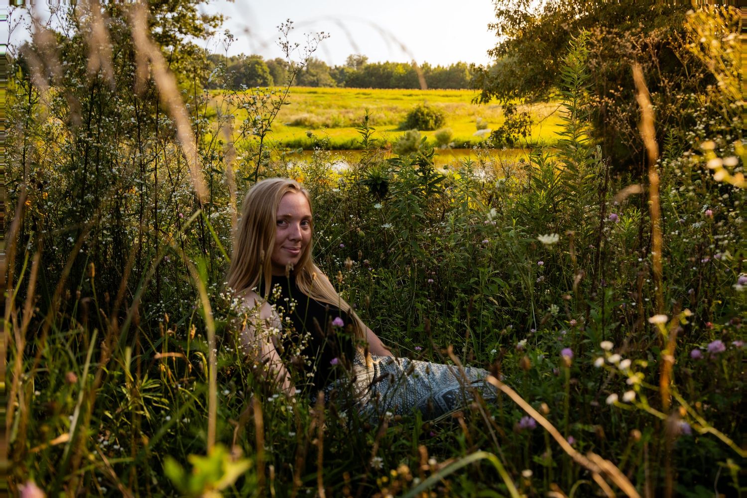 Young blonde woman sitting among wildflowers in a sunlit meadow with golden fields in the background.