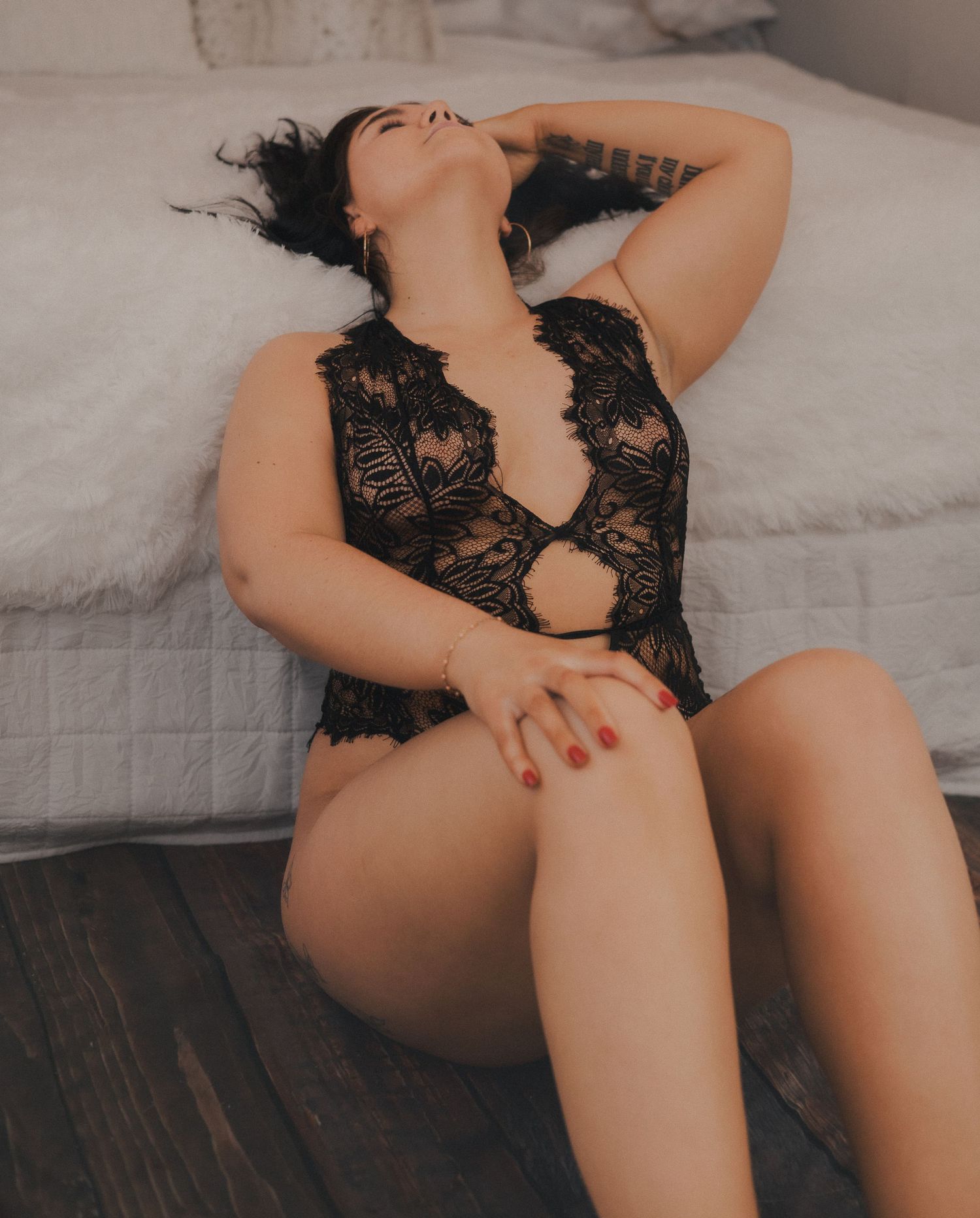 Woman in black lace bodysuit posing elegantly on floor beside bed in soft natural lighting.