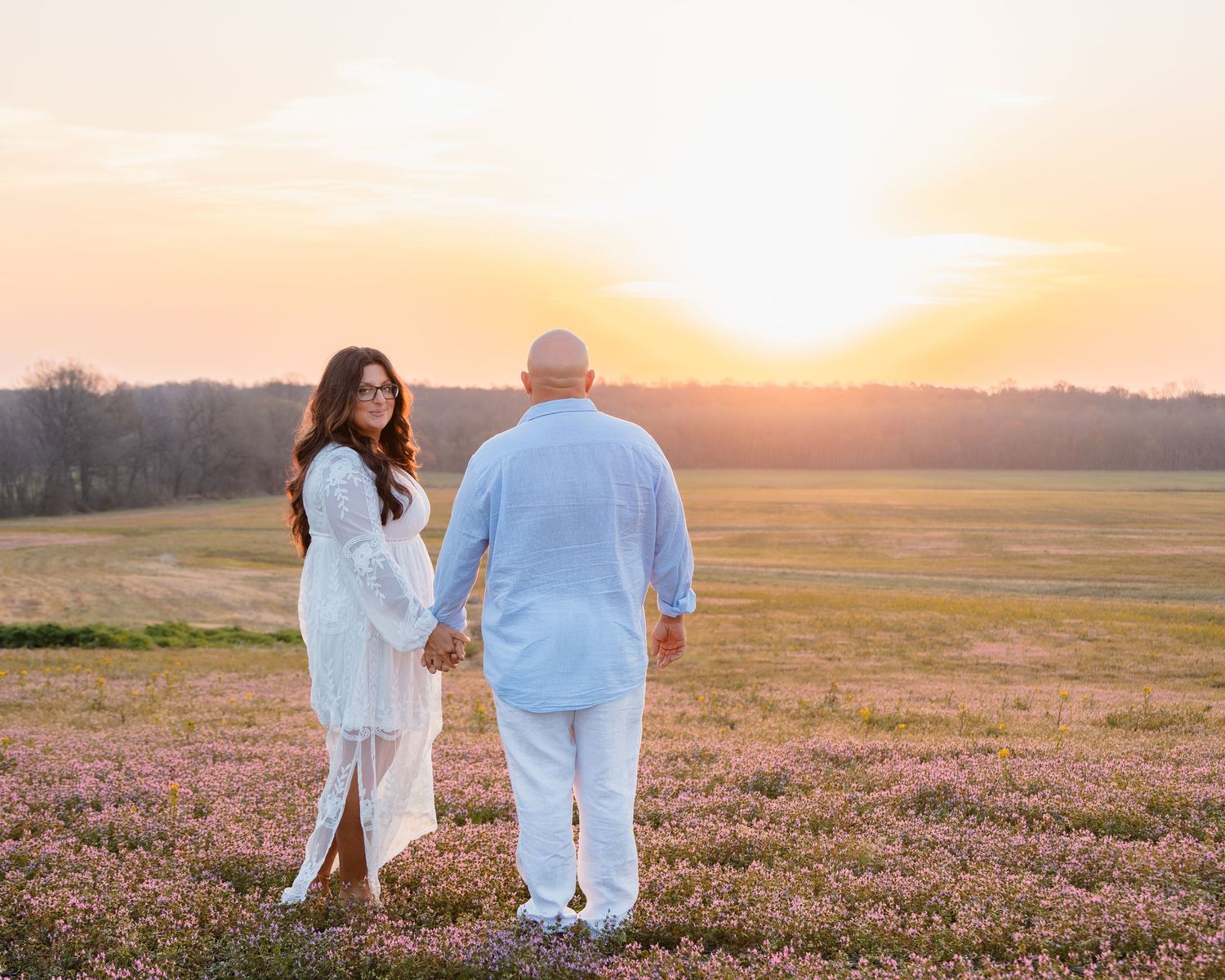 Couple holding hands in a blooming field at golden sunset, woman in white dress, man in light blue outfit.
