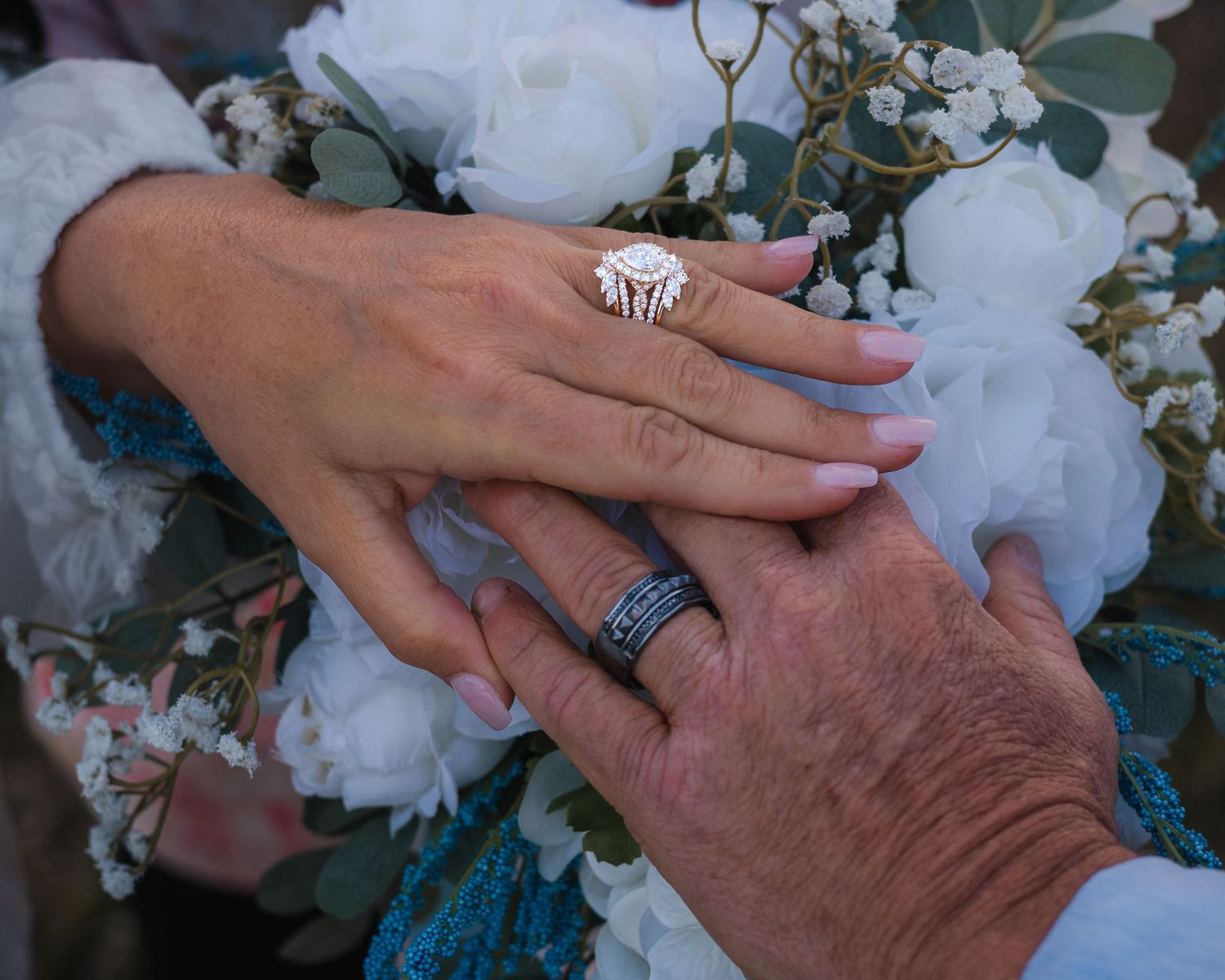 Close-up of bride and groom hands with diamond engagement ring and wedding band over white floral bouquet.