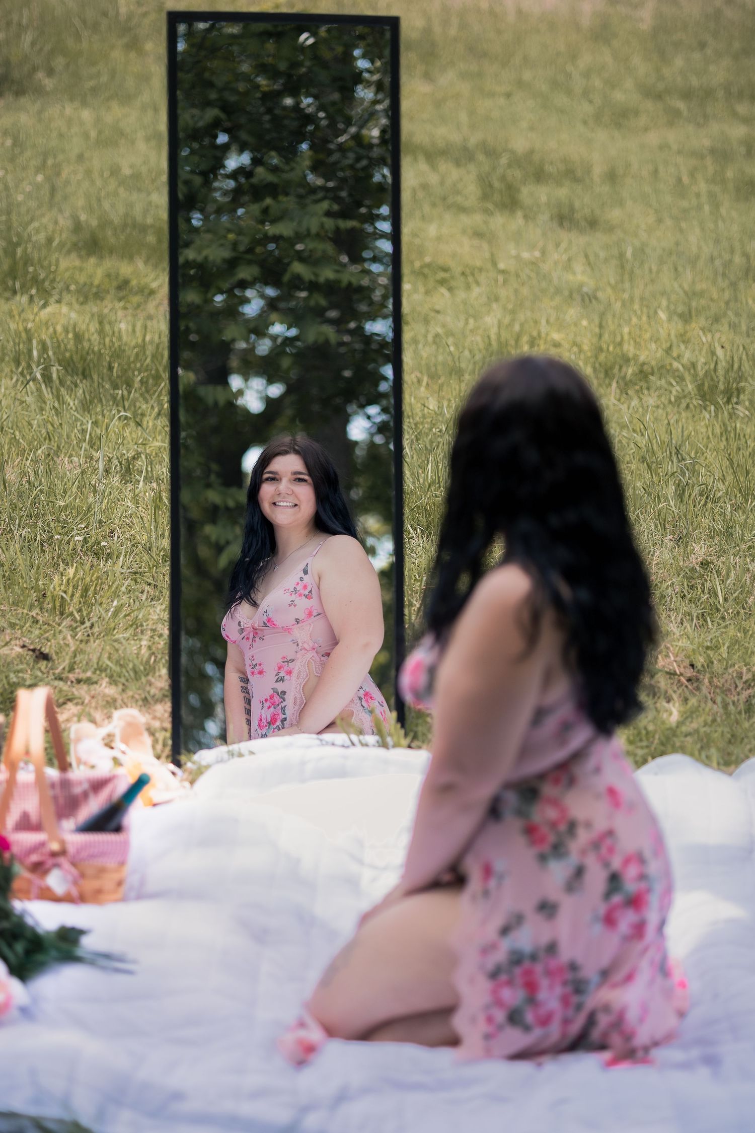Young woman in floral dress sitting on white blanket outdoors, smiling at her reflection in a tall mirror on green grass.